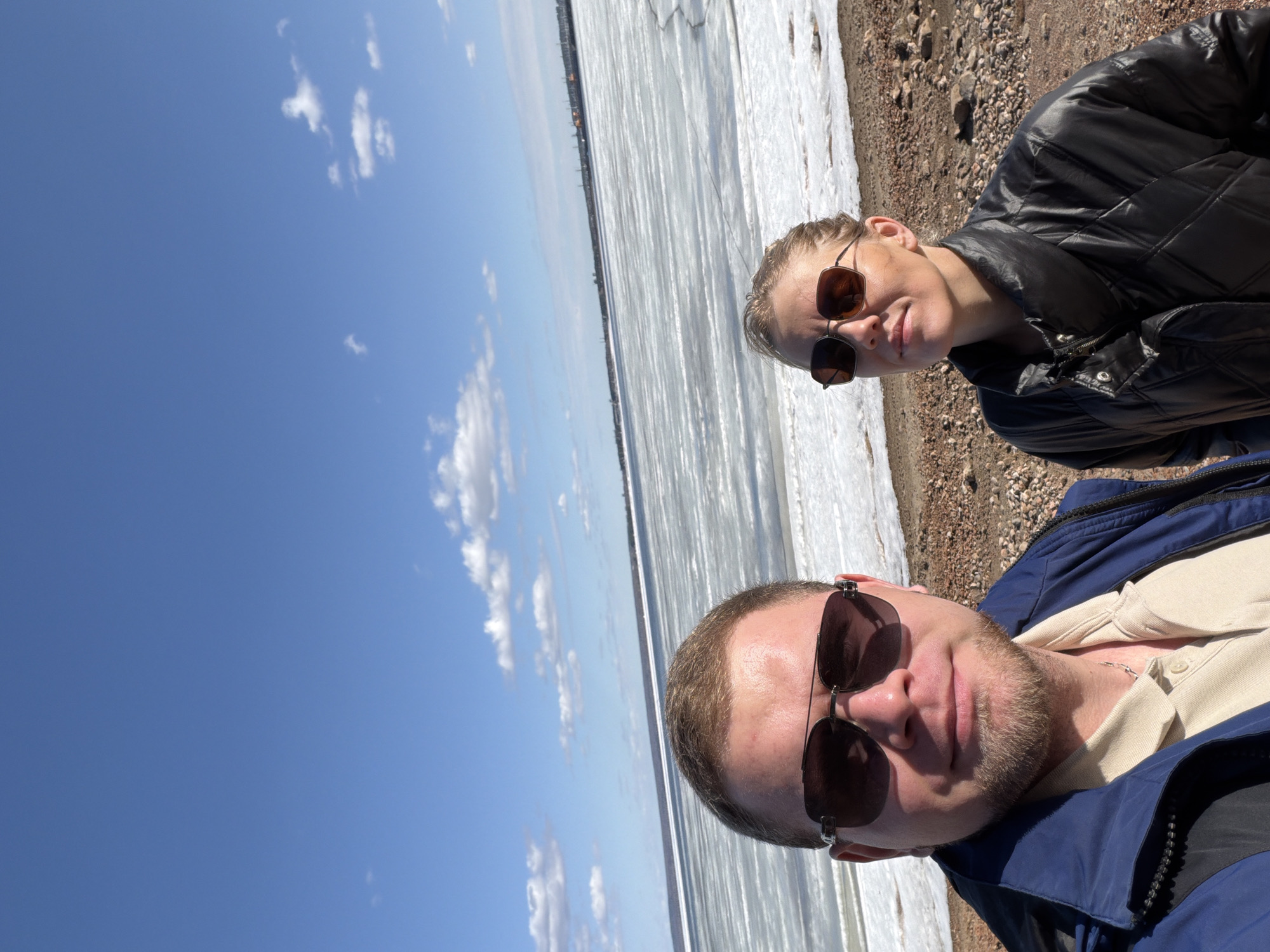 Two people, a male and a female, are posing for a selfie near a body of water covered in ice. The male is in the foreground, wearing sunglasses and a blue jacket over a light-colored collared shirt. The female is in the background, wearing sunglasses and a black puffy jacket. Behind them is a partially frozen body of water with a rocky shoreline. The sky is blue with scattered clouds. The picture was taken during the daytime, presumably in the city of Chaikovsky, Russia.