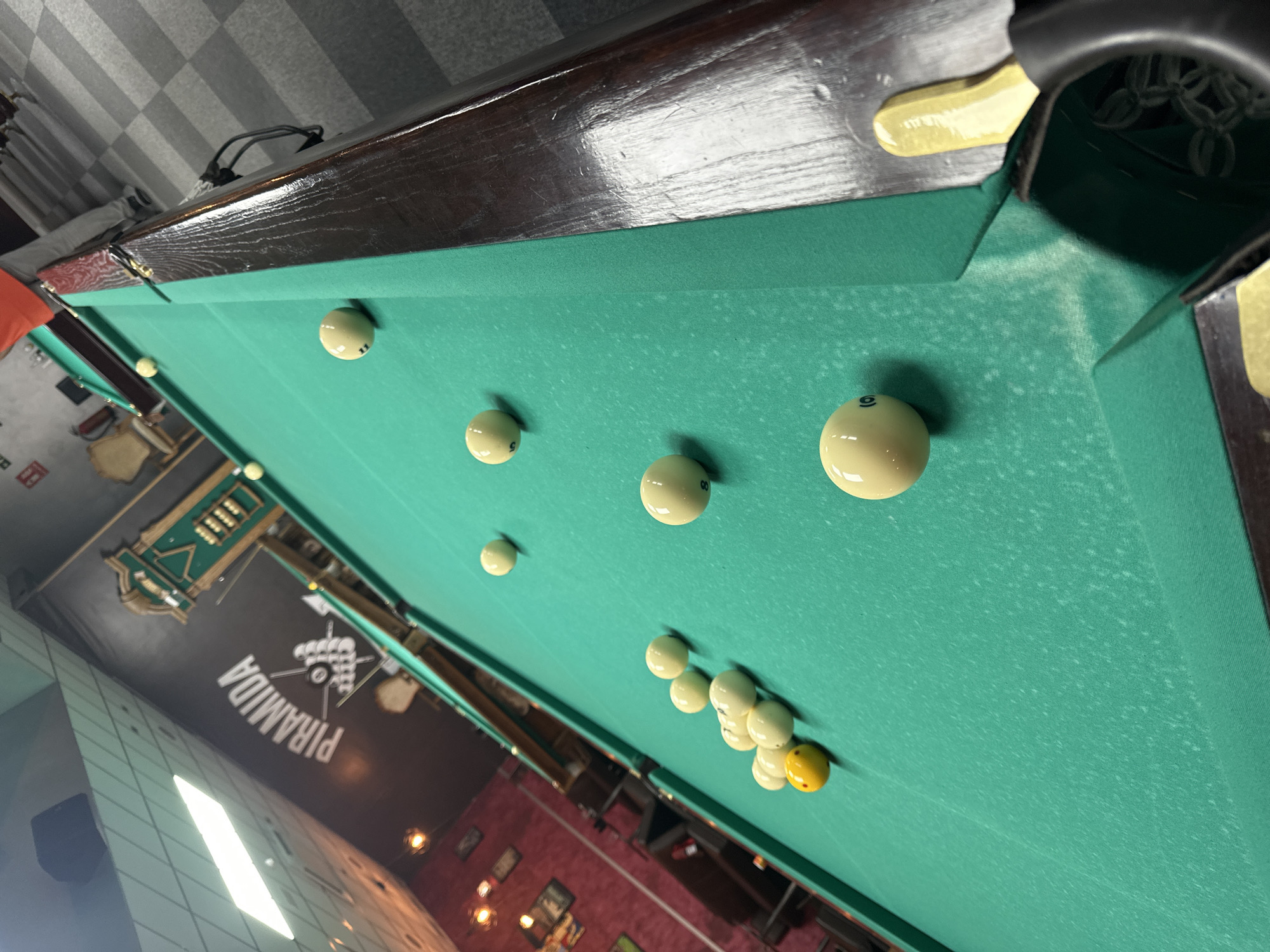 The image shows a pool table with billiard balls on a green felt surface. The table is surrounded by the interior of a establishment, visible through the skewed perspective, that features a sign with the word "PIRAMIDA" in Cyrillic alphabet. The lighting appears dim, and the decor includes visible wall decorations, possibly frames or pictures. The floor and ceiling are visible, suggesting the camera position is elevated and angled downwards at the table. I don't have information about location in the city of San Vicente Del Raspeig/sant Vicent Del Raspeig, Spain.