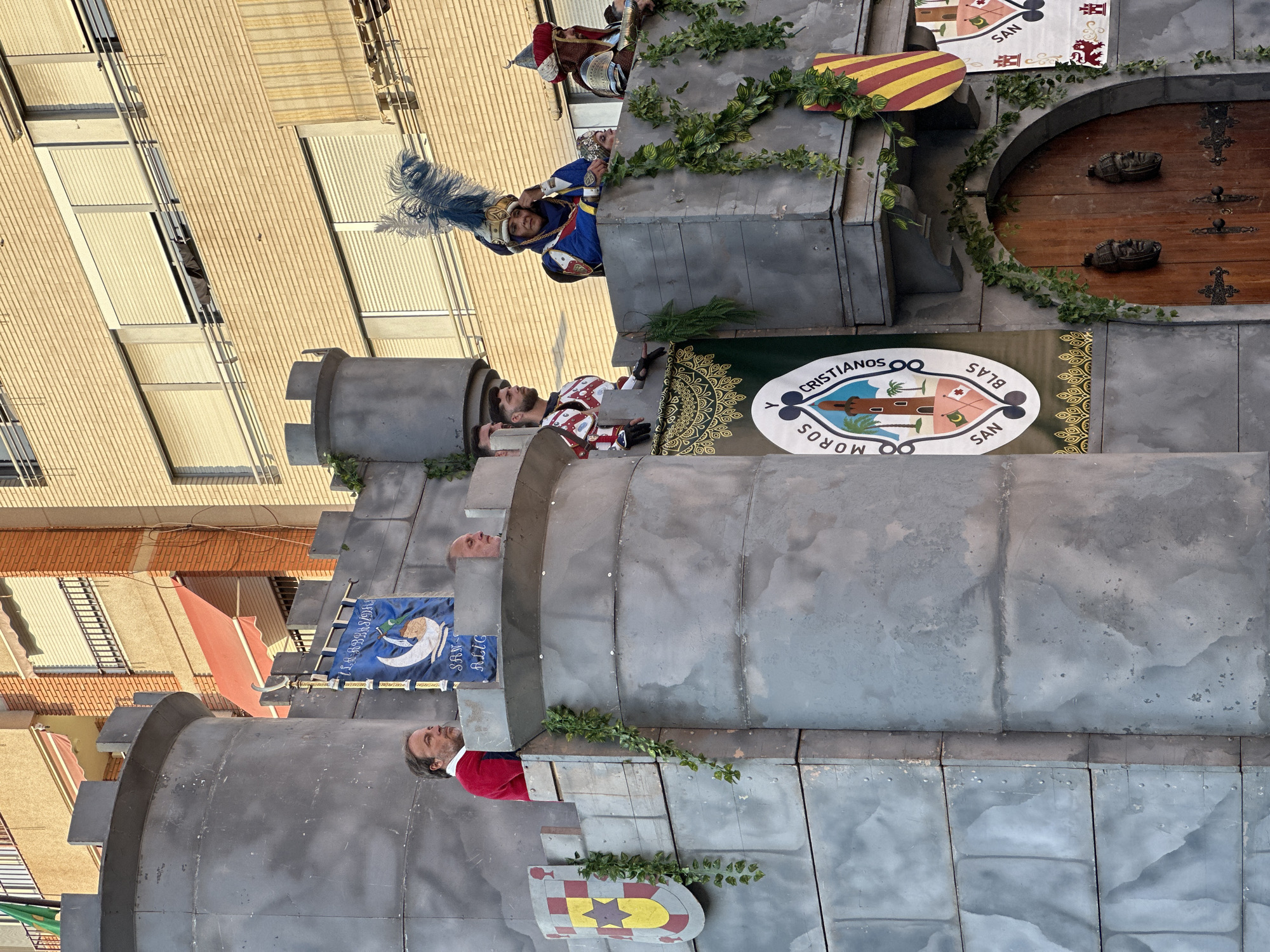 The image captures a scene from a parade, likely the "Moros y Cristianos" festival, in the city of Alicante, Spain. The backdrop is a large, grey, castle-like float, constructed from what appears to be painted cardboard or similar material, mimicking stone blocks. The float has ramparts and turrets, decorated with artificial ivy vines.
Several individuals are positioned on the ramparts, dressed in historical costumes. On the left, a man in a red jacket with white trim looks out, standing next to a flag depicting a crescent moon and a sailing ship. To his right, another man in a red and white medieval-style armor is visible. Further to the right, a person in elaborate blue and gold attire, wearing a large feathered headdress, appears to be speaking. Behind them, another figure in similar attire with a red turban is visible.
Banners are prominently displayed on the float. One large banner on the left features a shield with yellow and red stripes and a yellow star. Another prominent banner on the right, oval-shaped and white, displays the text "MOROS Y CRISTIANOS SAN BLAS" encircling an illustration of a tower with palm trees and flags. Smaller banners with similar motifs and heraldic symbols are also present. A large wooden door with decorative metalwork and carved faces is visible at the base of the float.
The float is positioned in front of buildings, suggesting an urban setting. The buildings are multi-story, with brick facades and windows covered by roller blinds. The lighting suggests it is daytime, possibly late afternoon due to the warm light. The overall impression is a festive and theatrical display, characteristic of cultural celebrations in Spanish cities.