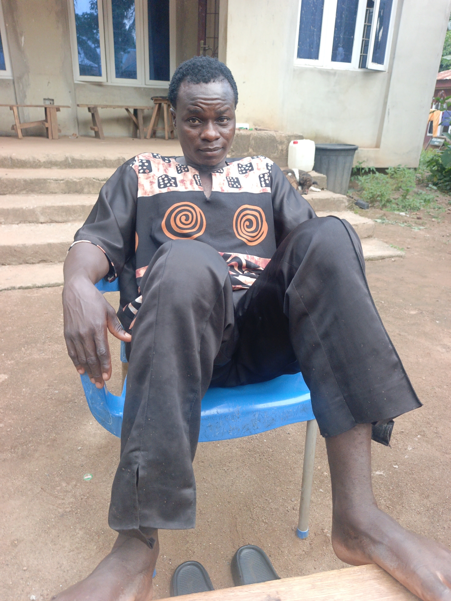 A man with dark skin and short, dark hair is sitting in a blue plastic chair. He is wearing a black shirt with an orange and white pattern and black pants. He is looking at the camera with a neutral expression. The background is a building with white trim around the windows. There are stairs leading up to the building and some vegetation. The ground is dirt. The scene appears to be outdoors, in a rural or semi-urban area. The building is likely a residential home or a small commercial establishment. The overall impression is one of a casual, everyday setting.