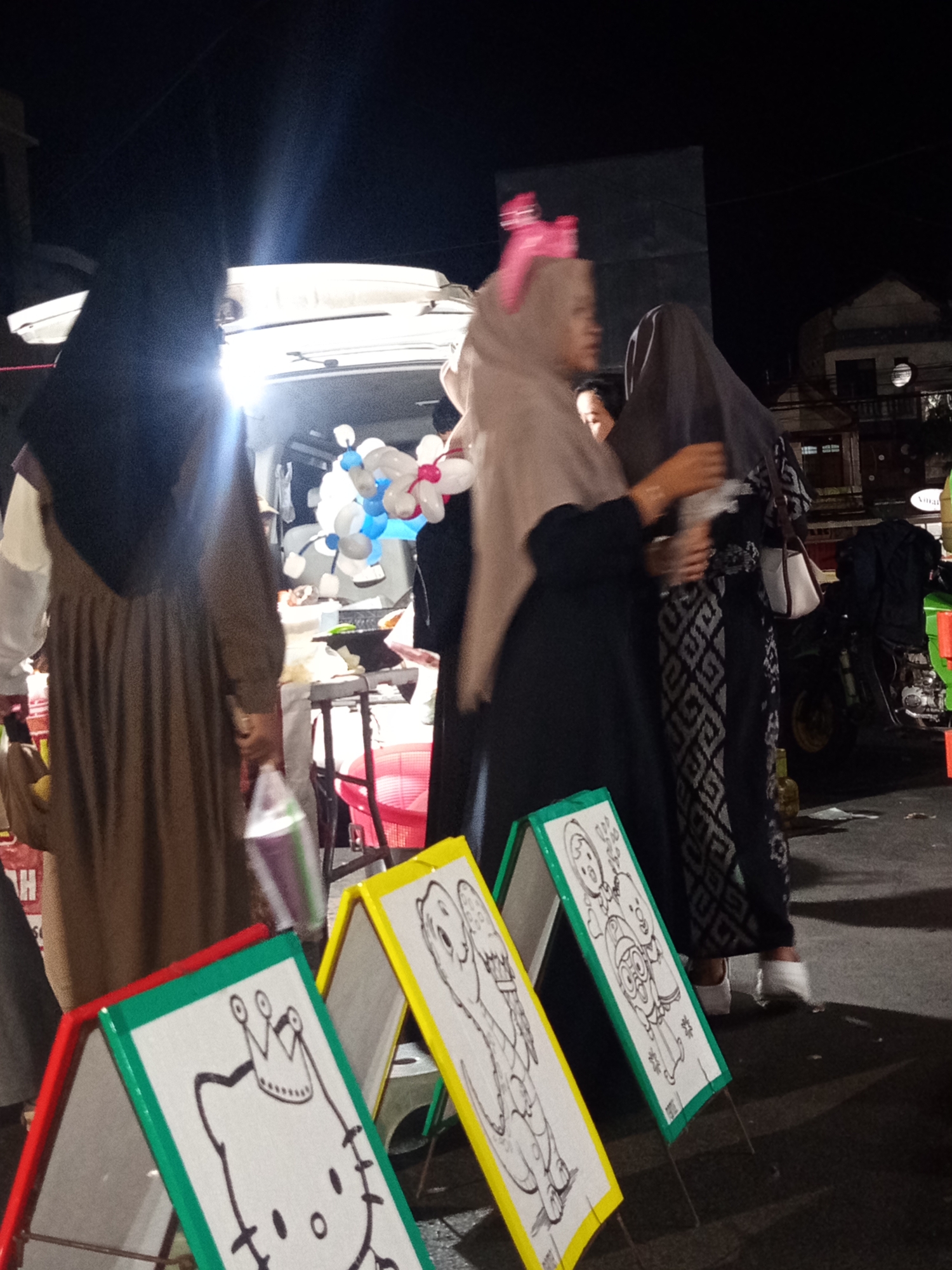 A nighttime scene depicts people gathered around a van with its rear doors open, suggesting a market or event. Several individuals, mostly women wearing hijabs, are visible. In the foreground, three A-frame drawing boards are set up, each displaying a line-drawn cartoon character, possibly for children's coloring activities. The boards are red, green, and yellow. In the background, there are balloons, a motorcycle, and buildings, indicating an urban setting. The overall impression is of a community gathering or a small business operating at night in Situbondo, Indonesia.