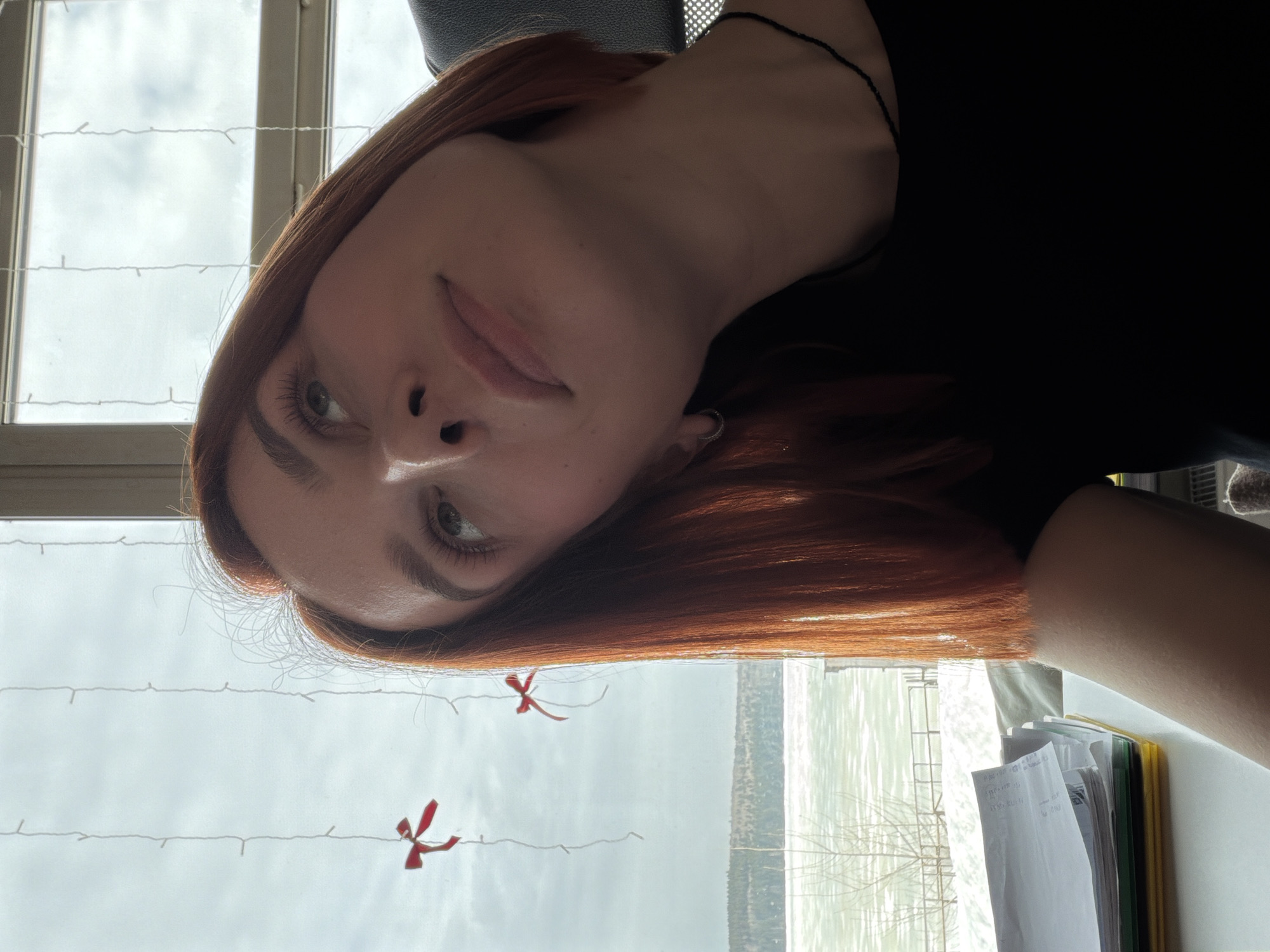 The image displays a close-up, angled portrait of a female subject, estimated to be between 20 and 30 years old, possessing shoulder-length reddish-brown hair and light-colored eyes. She is looking upwards and towards the viewer's right. The subject is situated indoors, facing a large window, wearing a black garment and a thin black beaded necklace. A silver hoop earring is visible in her left ear. Through the window, an overcast sky and a winter landscape are visible. A significant body of water, consistent with the Votkinsk Reservoir on the Kama River in Chaikovsky, Russia, is partially covered by ice and snow. The immediate shoreline features bare deciduous trees, with a dense coniferous forest visible on the distant bank. Two vertical strands of white string, each adorned with a small red bow, are suspended in front of the window glass. In the lower-left foreground, a stack of white papers rests on a yellow folder on a flat, light-colored surface, indicative of a desk. A portion of a black chair with a visible mesh back is present behind the subject's right shoulder.