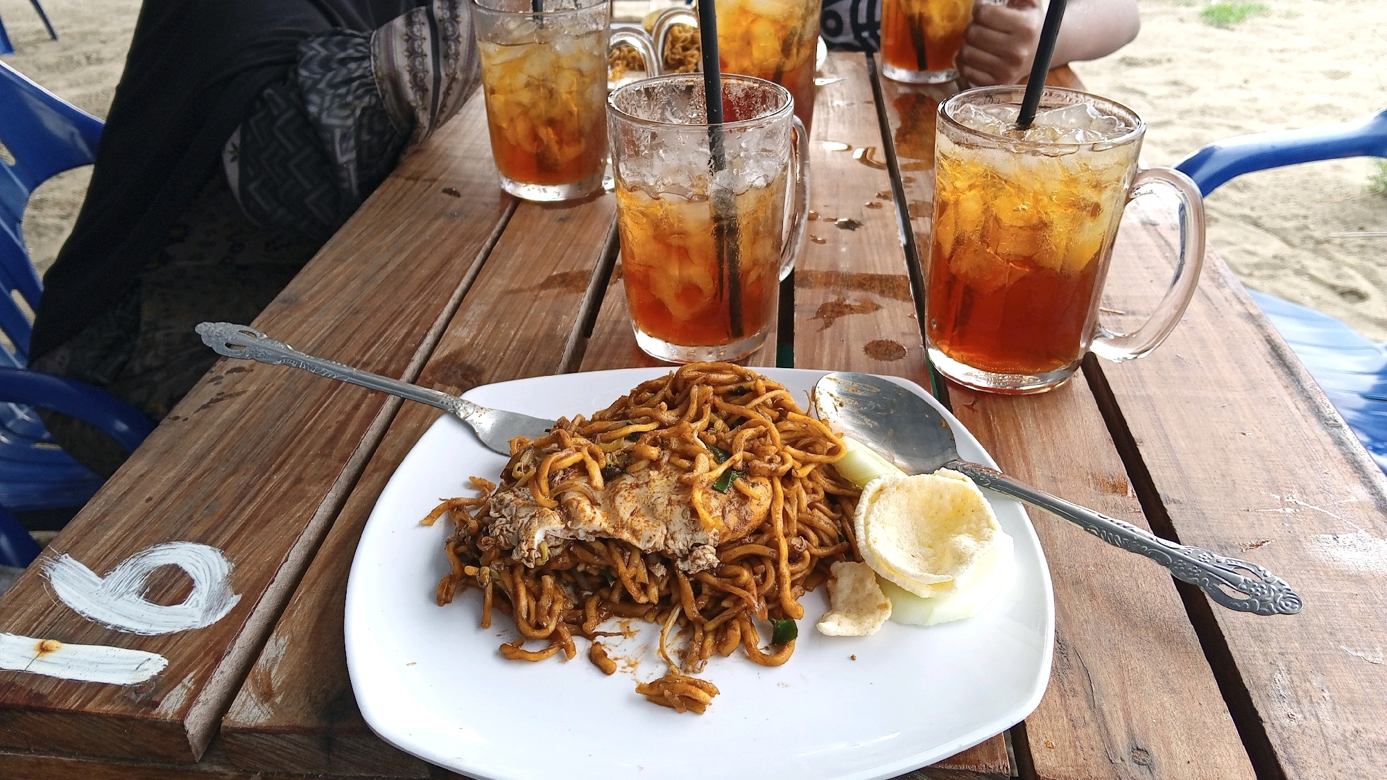 A plate of fried noodles is served on a wooden table next to three glasses of iced tea. The noodles are brown and appear to be mixed with vegetables and egg. There are also some crackers and cucumber slices on the plate. The iced tea is in clear glasses with straws. The table is outdoors, and there is sand and a blue chair visible in the background. The setting appears to be a casual dining area, possibly a beachside cafe in Kruengjuli Timu, Indonesia, given the sandy environment and the style of the refreshments and meal. The image is captured from a slightly elevated angle, focusing on the food and drinks.