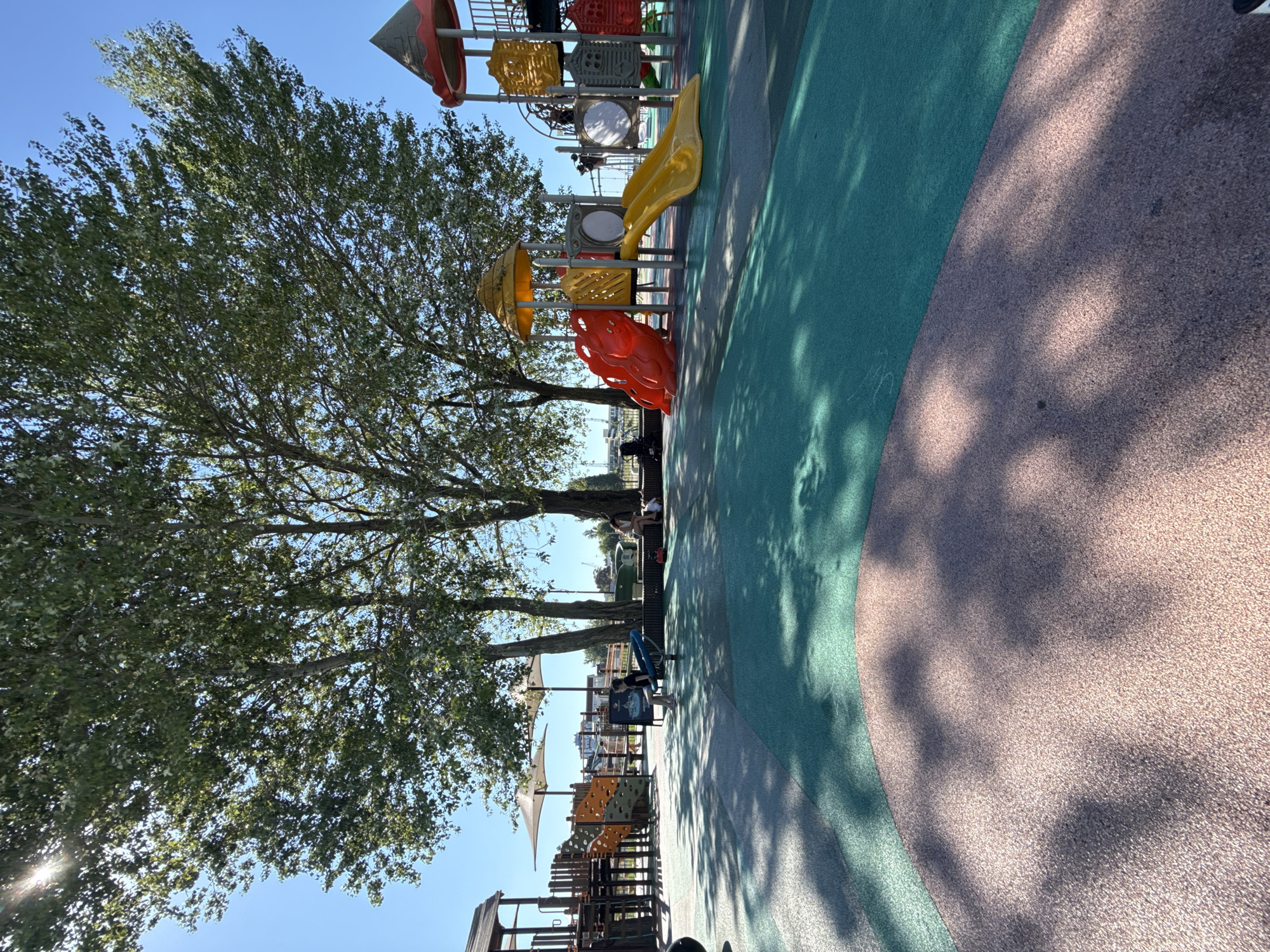 The image depicts a modern, colorful playground with a soft, multi-colored rubberized surface. In the background, large, leafy trees cast dappled shadows across the play area. A bright blue sky dominates the upper portion of the frame.
The playground features several structures, including a bright red, multi-level climbing structure with yellow slides. To the left, a wooden play fort with orange and green climbing elements is visible. In the middle ground, a child is seated on a blue seesaw-like apparatus, appearing to be alone. Further to the right, a group of people, including a woman and a child, are seated on a wooden bench beneath the shade of the trees. In the far background, there are hints of urban structures and possibly water bodies, suggesting an open, potentially recreational area within a city.
The overall scene is one of a sunny day at a public park or playground, likely in a temperate climate given the lush green foliage of the trees. The presence of well-maintained play equipment and recreational seating indicates a deliberate design for public use. The lighting suggests it is daytime, with the sun appearing high in the sky, creating strong shadows.