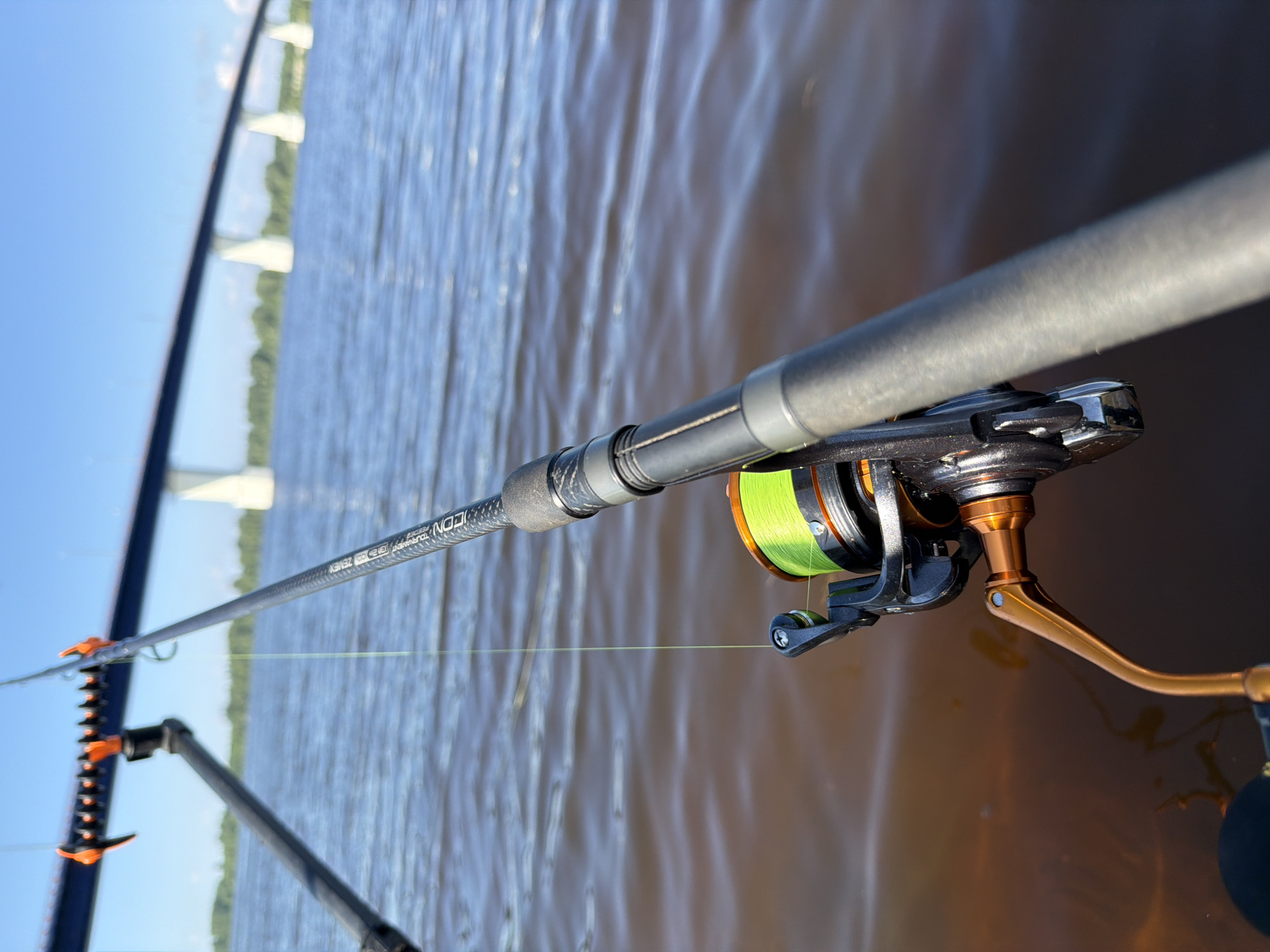 A close-up, eye-level, angled outdoor shot focuses on a fishing rod and reel positioned over murky brown water. The rod, made of a textured dark material with grey sections, extends from the right side of the frame towards the center. A fishing reel, featuring a neon green line wound around its spool, is attached to the rod. The reel has black and orange accents, and a golden handle is visible. The water's surface is rippled, suggesting a slight breeze. In the background, a wide, modern bridge with white concrete pillars spans across the water under a bright blue sky. Green trees line the distant shore. The scene appears to be a calm day for fishing. While the specific location of Tarasovo, Russia, cannot be confirmed from the visual information alone, the presence of a bridge and a body of water are evident. No people are visible in the image.