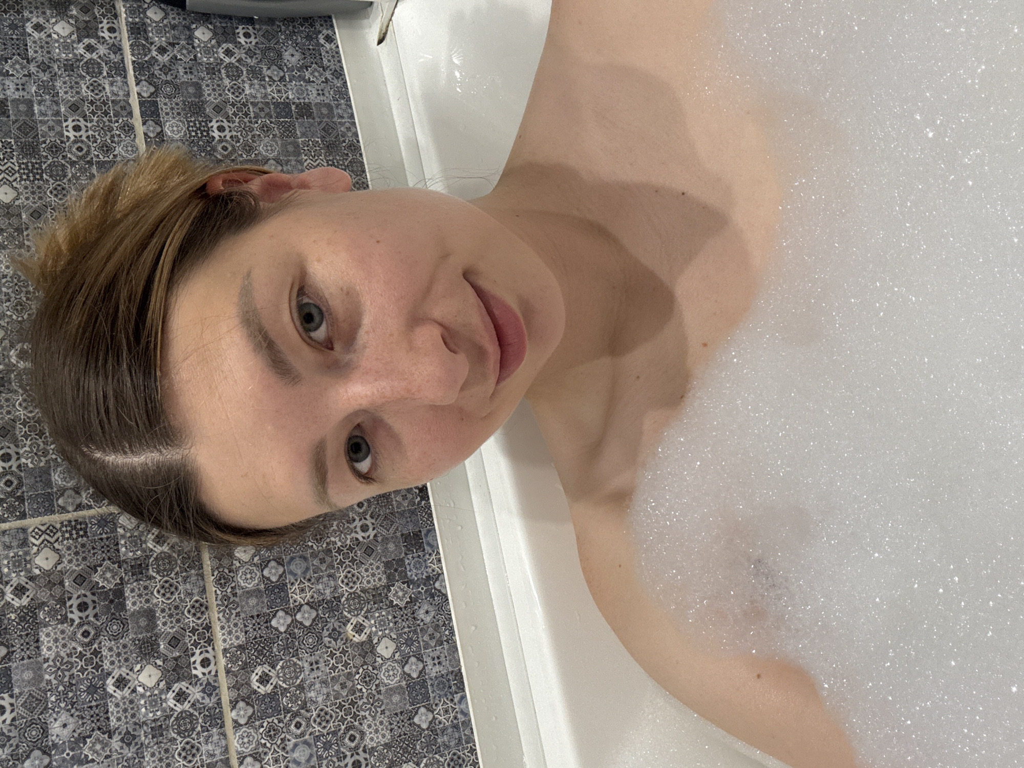 A woman is lying in a bathtub filled with bubbles. Her head is near the edge of the tub, close to a wall covered in patterned blue and grey tiles. The woman is looking at the camera. The location cannot be determined from the image.