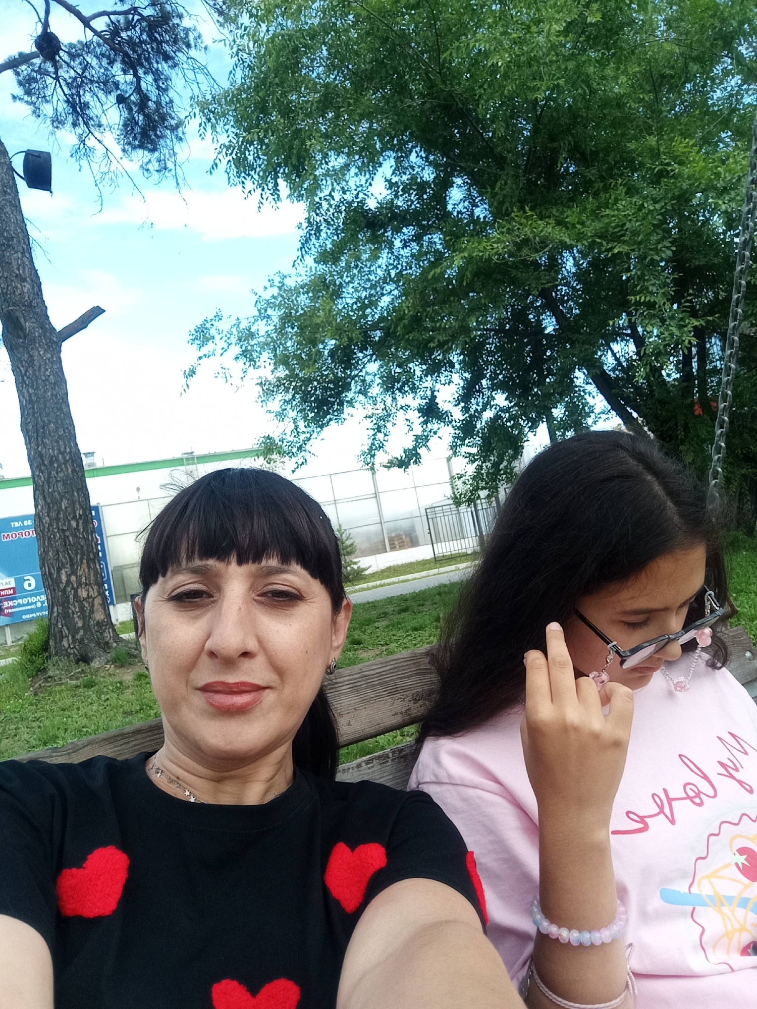 In Belogorsk, Russia, two females, an adult and a child, are seated on a wooden bench outdoors. The adult, positioned on the left, has dark hair with bangs and is wearing a black t-shirt adorned with red hearts. The child, on the right, has long dark hair and is wearing a pink t-shirt, glasses with pink flower accents, and a multi-colored bracelet. They appear to be on a swing set, with the chain of the swing visible on the right side of the frame. In the background, a large tree stands to the left of the subjects, and lush green foliage and a bright blue sky are visible overhead. A building with large windows and a fence is also present in the background. The ground around the bench is covered in grass.