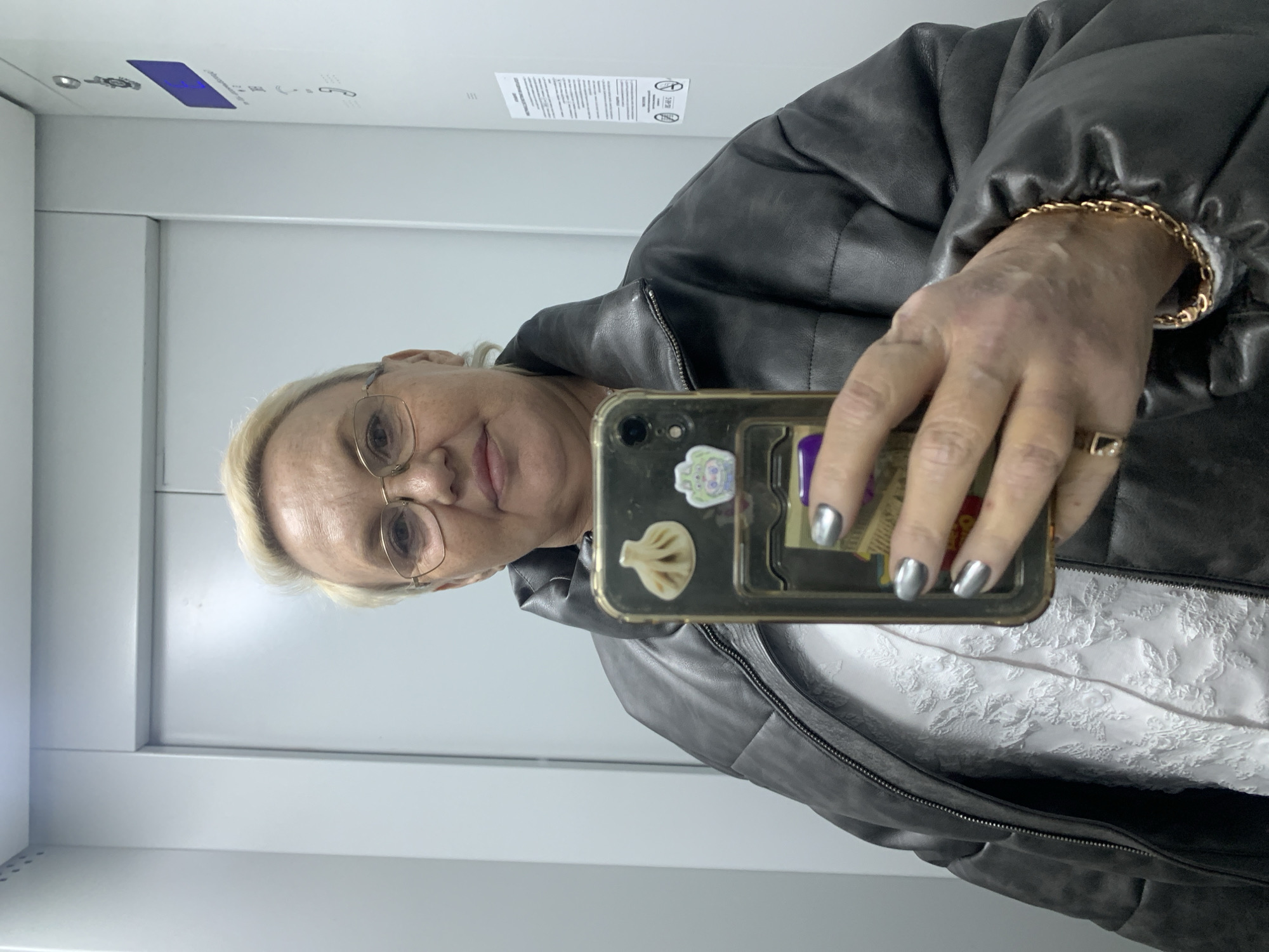 The image shows a woman in what appears to be an elevator. She is holding a smartphone with stickers on the back, using it to take a selfie. She is wearing glasses, a black puffy jacket, and a gold bracelet. The elevator interior is white with a panel on the ceiling displaying information in Russian, suggesting the location is likely in a Russian-speaking country. Based on the metadata, the location is Chaikovsky, Russia.