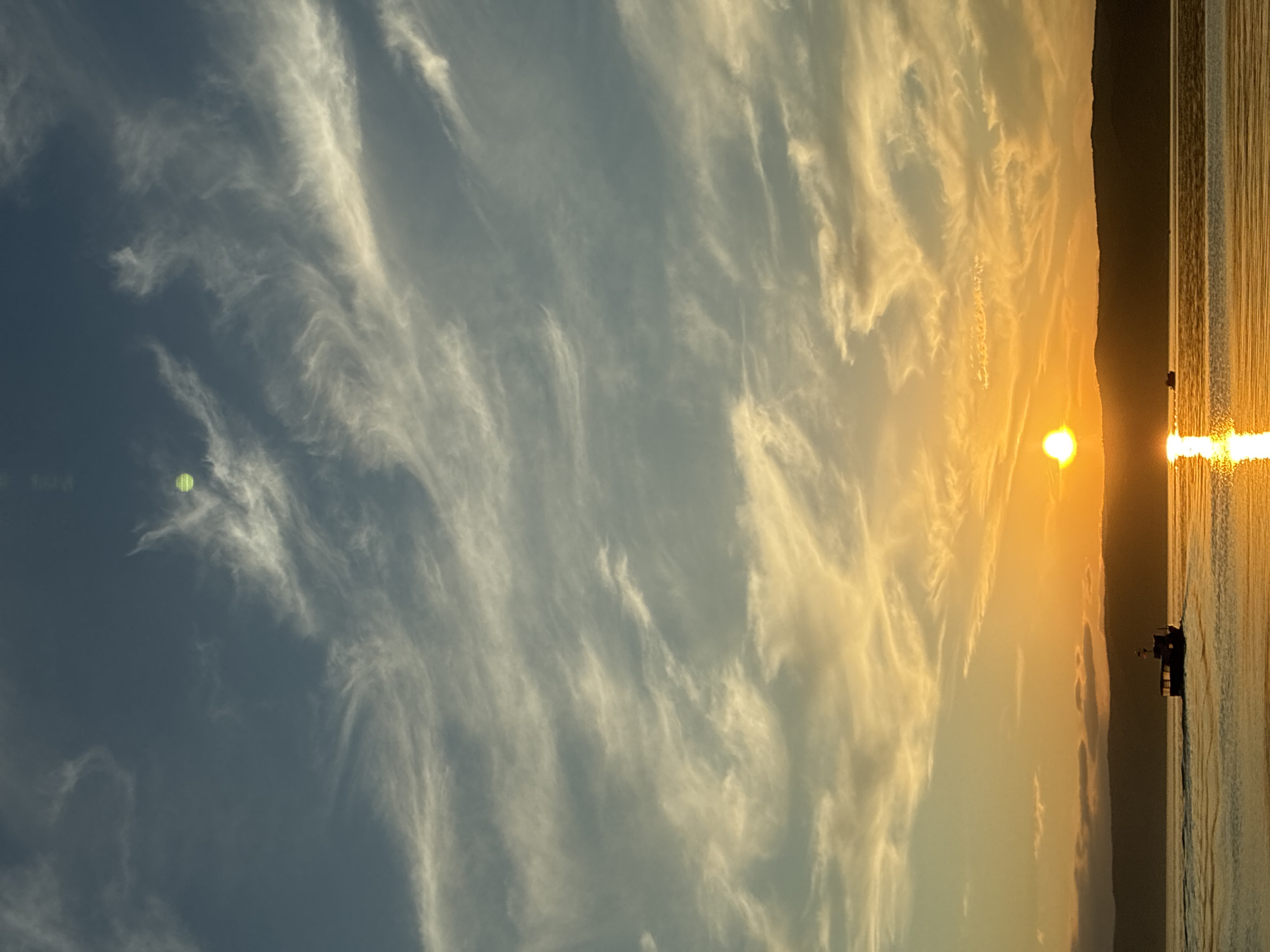 The image captures a serene sunset over Lake Baikal, viewed from the shore in Listvyanka, Russia. The sky is a gradient of deep blue at the top, transitioning to soft yellows and oranges near the horizon, with wispy white clouds scattered throughout. The sun, a bright orb, is low on the horizon, casting a shimmering golden reflection on the calm water. A small boat is visible in the middle ground, moving from left to right across the lake, leaving a faint wake. In the background, a dark, silhouetted mountain range forms the distant shore. The overall atmosphere is peaceful and tranquil, characteristic of a summer evening in Listvyanka.