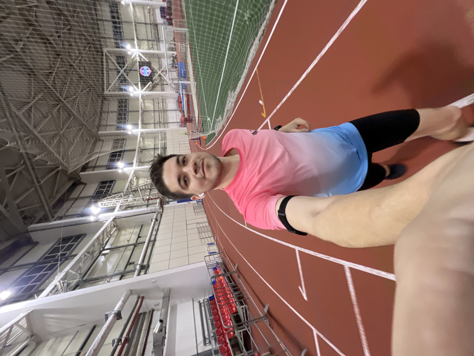 The image shows a man taking a selfie indoors at what appears to be an athletic facility, specifically a running track. The photograph is oriented with the man lying on his back, looking up, and his body positioned diagonally across the frame, suggesting it was taken from a selfie stick or outstretched arm held at an angle and then rotated, or perhaps a camera mounted on his body.
The man, who appears to be in his 20s or 30s, is smiling faintly and looking directly at the camera. He has short, dark hair and is wearing a long-sleeved athletic shirt that features an ombre design, transitioning from pink at the top to light blue at the bottom. He is also wearing dark, possibly black, athletic shorts or leggings. A dark-colored smartwatch or fitness tracker is visible on his left wrist, and a white earbud is in his right ear. His right arm, which is holding the camera, is partially visible in the bottom right corner of the frame.
The background reveals a large indoor sports arena. The dominant feature is the reddish-brown running track with white lane markings. To the left of the man (or above him in the rotated image), there's a green artificial turf field, partially visible, with white netting separating it from the track area. A white soccer goal is also visible on the field.
Further in the background, a large, intricate ceiling structure made of metal trusses and supports is visible, typical of large indoor sports complexes. Numerous bright lights illuminate the facility. On the far side of the track, there are tiered red seats, indicating spectator stands. High above the track and field, a digital display or scoreboard is visible, showing a circular graphic with multiple colors, though specific details or text are indistinguishable. The overall impression is a modern, well-lit indoor sports complex where the man is likely engaged in or preparing for physical activity.