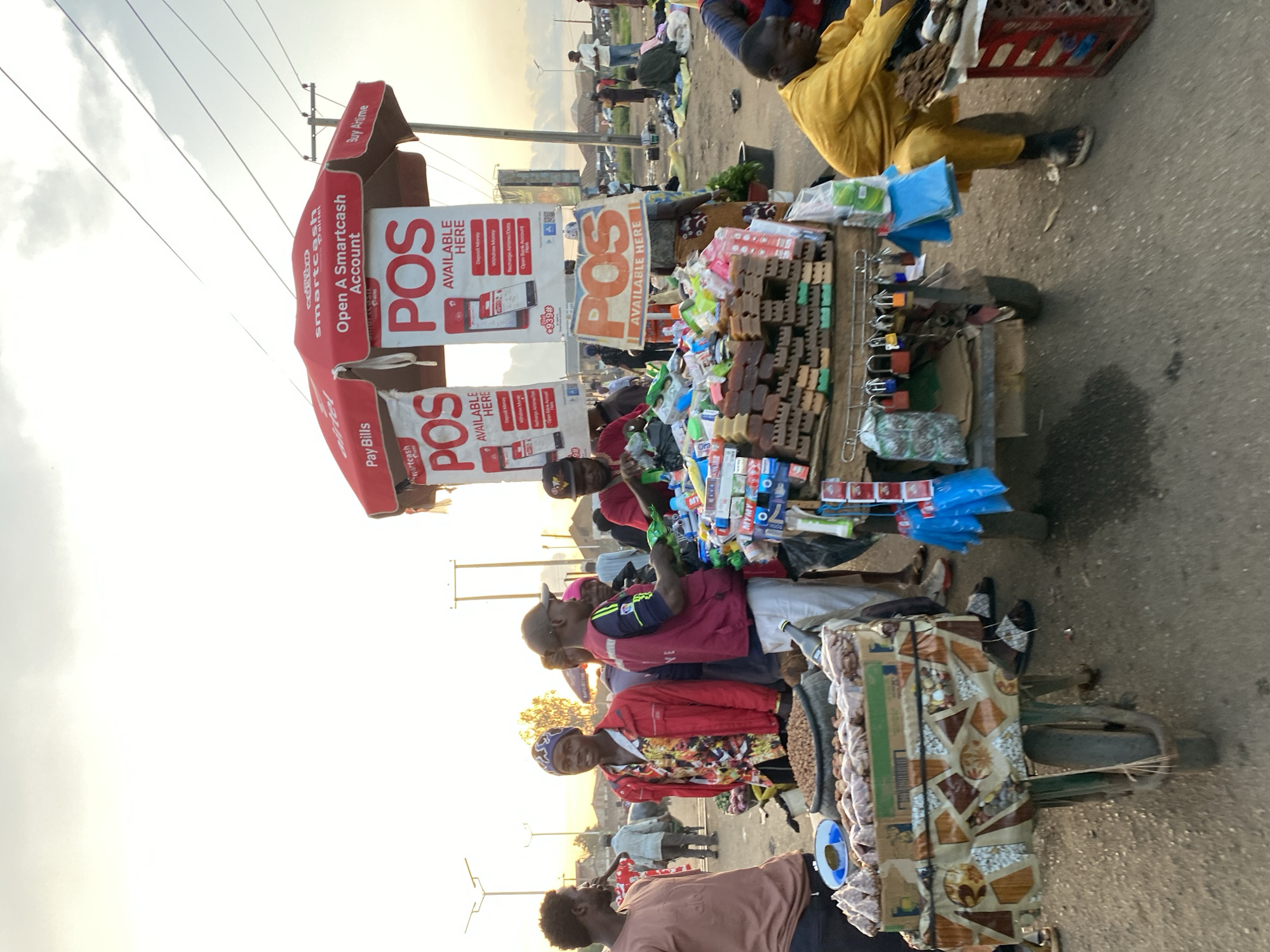This image captures a bustling outdoor market scene in Bukuru, Nigeria, during late afternoon or early evening. In the foreground, two vendor carts display various goods.
The central feature is a red promotional umbrella advertising "Airtel smartcash" with slogans like "Open A Smartcash Account," "Pay Bills," and "Buy Airtime." Three white banners hanging from the umbrella prominently display "POS AVAILABLE HERE," accompanied by illustrations of a smartphone app showing options for "Deposit Money," "Withdraw Money," "Recharge Airtime/Data," and "Open Bank Account Here," along with the USSD code "*939#". A smaller, separate sign also states "POS AVAILABLE HERE."
Directly beneath the umbrella, a multi-tiered vendor cart is laden with merchandise. The top tier holds an assortment of colorful packaged goods, including snacks, toiletries, and small plastic items. Below this, rectangular brown bricks, consistent with packaged soap or similar products, are stacked, with green items visible underneath. The bottom section of this cart features hooks displaying multiple colorful padlocks and blue plastic bags. A man in a dark vest and baseball cap stands behind this cart, reaching towards items, while another man in a red t-shirt and cap is actively arranging green plastic bags.
To the left of this main cart, a green wheelbarrow-like cart, adorned with patterned fabric, holds a large tire filled with peanuts and several small packages of what appear to be nuts or snacks. A blue and white plate is placed on this cart. A man in a brown t-shirt stands to the far left, observing the scene, while another man in a red jacket, colorful patterned shirt, and blue beanie stands nearby, looking forward.
In the lower right, a man in a yellow outfit is seated on a low stool, attending to items on a red plastic crate. The ground is a mix of worn pavement and dirt. In the mid-ground and background, numerous indistinct figures suggest a busy market environment. Power lines crisscross the overcast sky, and distant buildings are visible beneath the horizon, illuminated by diffused, warm light. Utility poles are present throughout the scene.