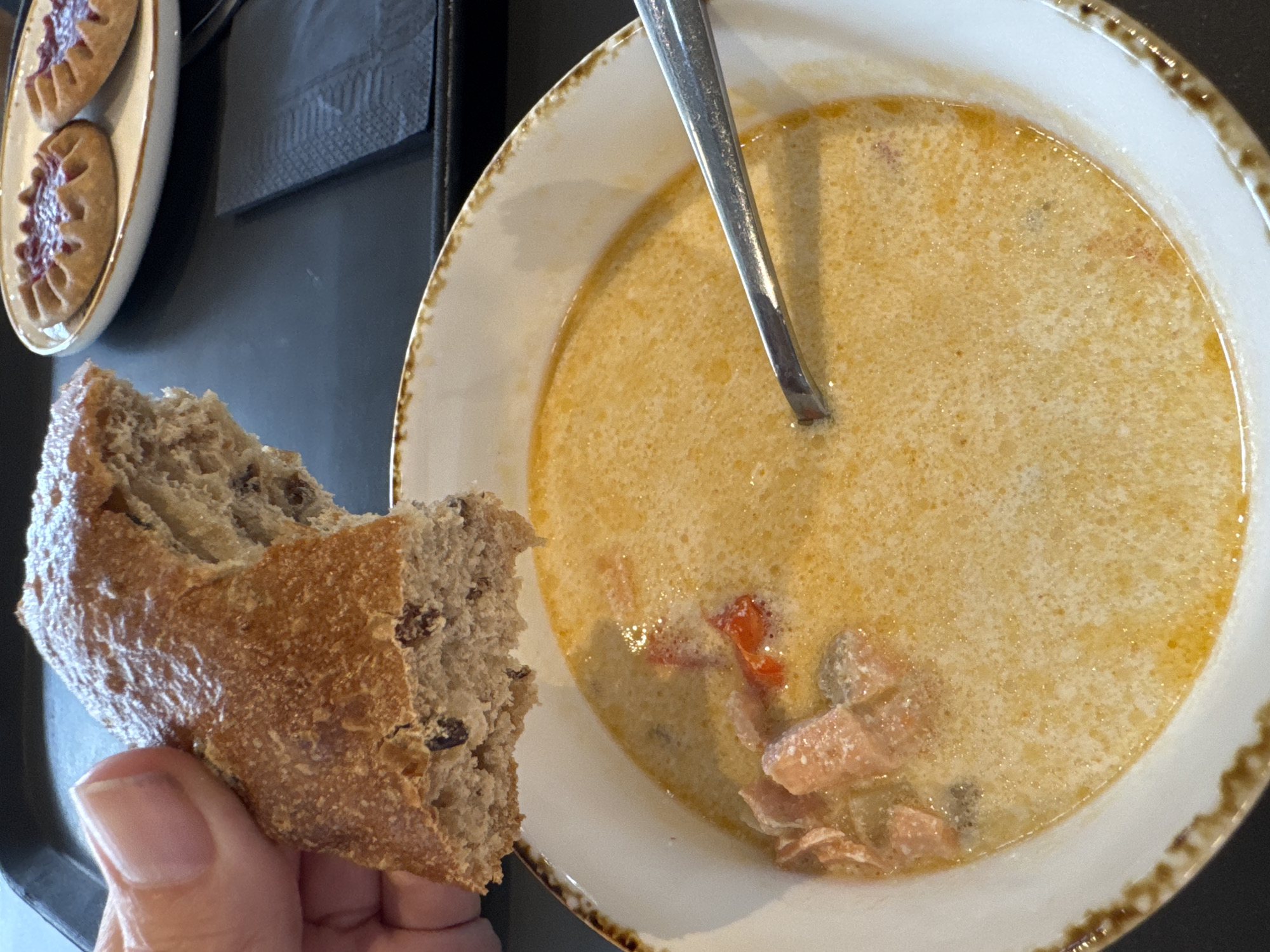 A close-up shot captures a piece of dark, rustic bread being held by a person's thumb and index finger over a bowl of creamy salmon soup. The bread has visible raisins. The soup contains chunks of salmon and red peppers. A silver spoon rests in the soup. In the background, a small white plate holds two small, round pastries topped with a red jam, typical of Karelian pies, a delicacy associated with the city of Lakhdenpokhya, Russia. The setting appears to be a casual dining environment, possibly a cafe or restaurant.