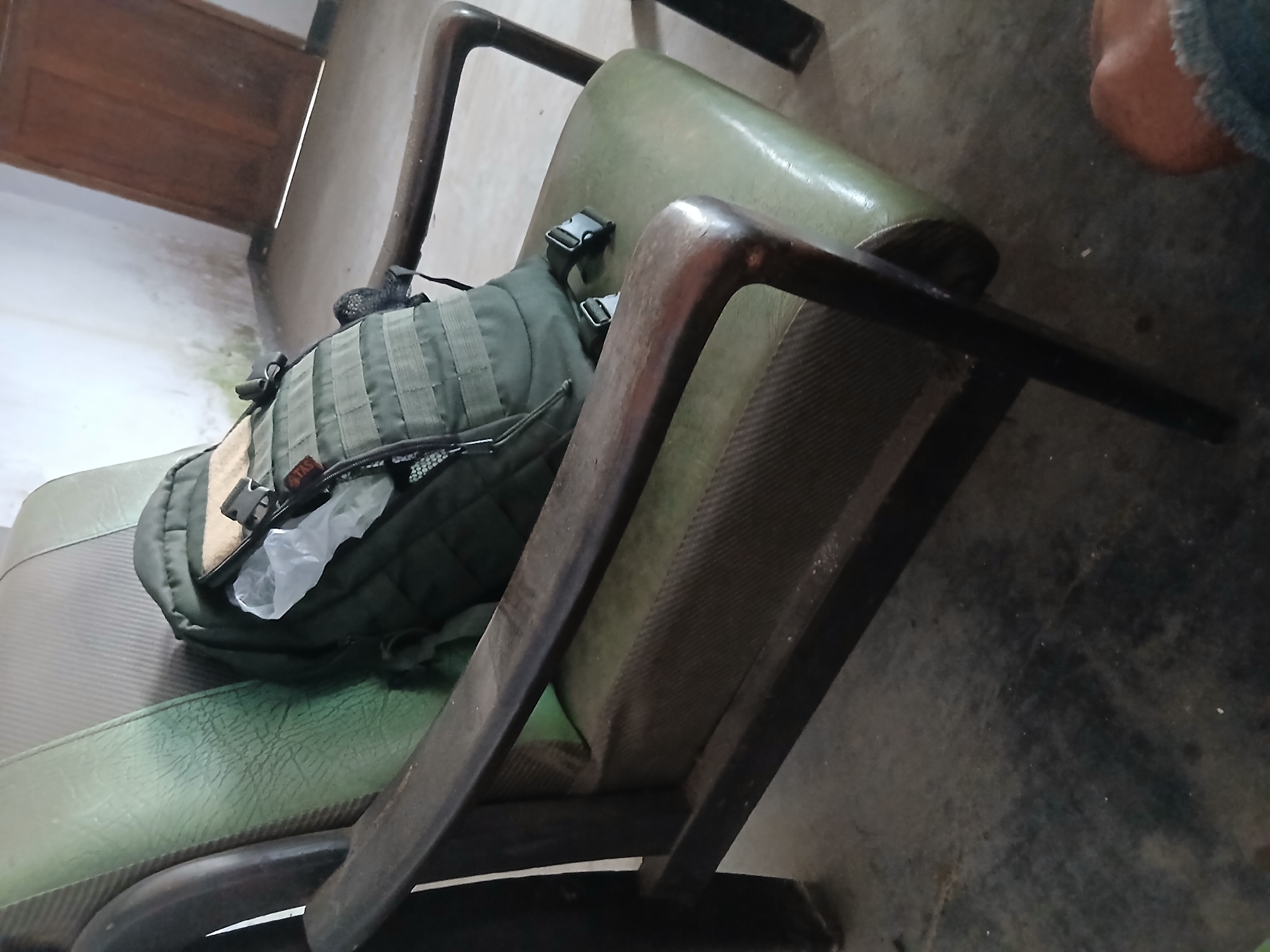 A green backpack is placed on a worn, green upholstered armchair in a sparsely furnished room. The backpack appears to be a tactical style, with MOLLE webbing visible. The armchair has dark wooden arms and legs. The room has concrete flooring and white walls with some water staining. A wooden door is visible in the background. The scene is likely within a residence in Kademangan Kulon, Indonesia, given the context.