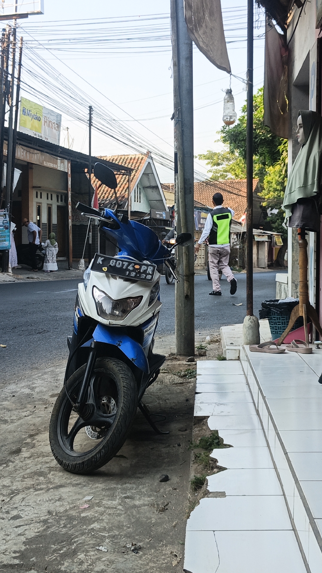 The image shows a street scene in Dawe, Indonesia. In the foreground, a blue and white motorcycle is parked on the side of the road. A person wearing a green safety vest is walking away from the camera down the street. The street is lined with buildings and utility poles with numerous overhead wires. A mannequin dressed in a hijab is visible on the right side of the frame. The overall scene depicts a typical urban street environment.