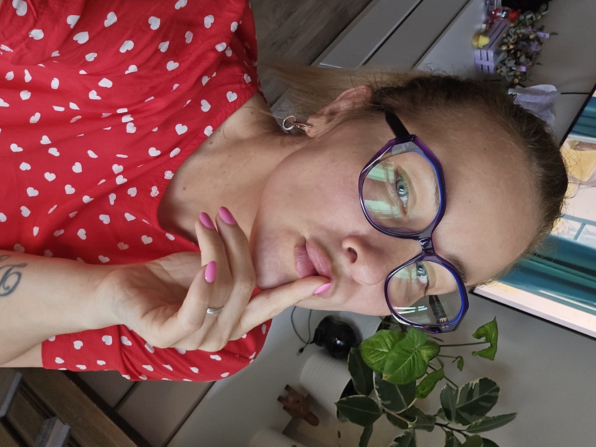 A woman is visible in the image, wearing a red polka dot top and eyeglasses. She is holding her finger to her lips. Her fingernails are painted pink. She also has a ring on her finger and a tattoo is visible on her wrist. A green plant is in the lower right corner of the frame. There is a table in the background with some objects on it, as well as a computer screen. I cannot confirm that the location is Sochi, Russia based on the image.