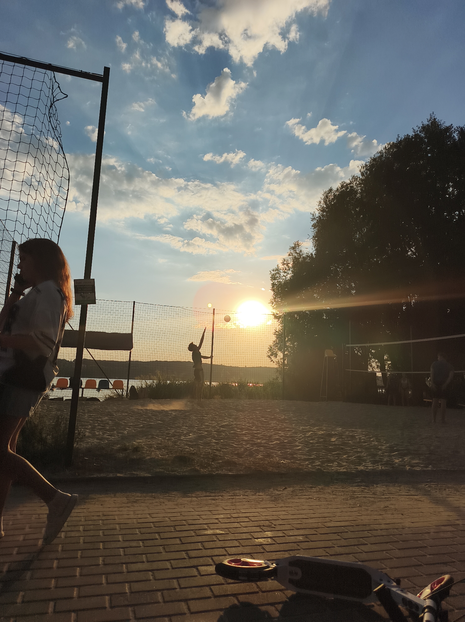 The image depicts a scene at a beach volleyball court, likely during sunset, located in Ternopil, Ukraine. In the foreground, a person with red hair is walking from left to right, partially obscured by a volleyball net. The court itself is made of sand, with a net dividing it. Two figures are visible on the court, one in the process of hitting a volleyball, and another standing further back. Beyond the court, a body of water and a distant shoreline are visible. The sky is partly cloudy with the sun setting, casting a warm glow over the scene and creating lens flare. In the lower right corner of the image, a scooter lies on a paved pathway.