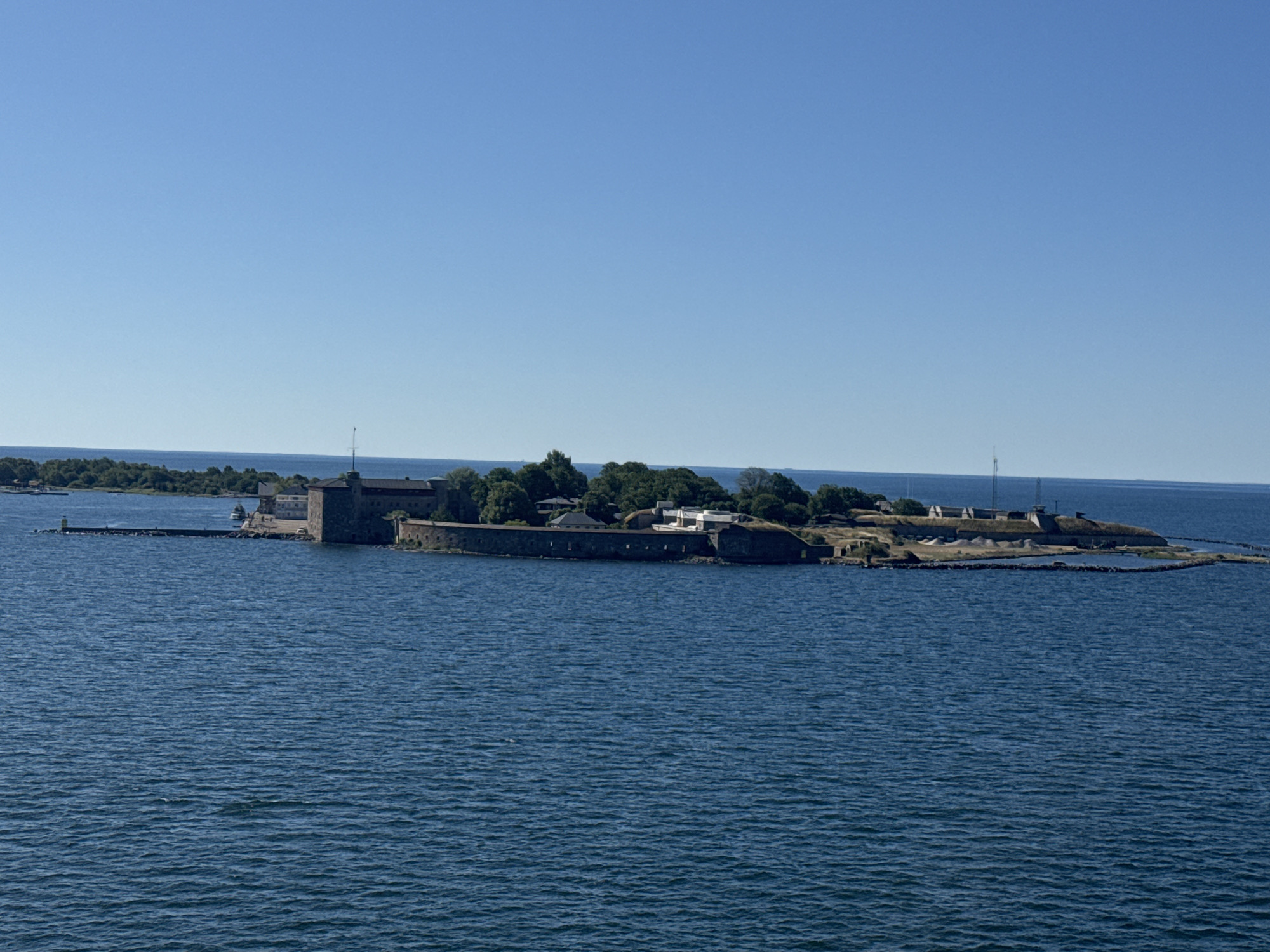The image displays a wide-angle, eye-level view of the island fortress of Kungsholmen in Karlskrona, Sweden, on a clear, sunny day. The fortress, characterized by its robust stone walls and buildings, is situated on a strip of land extending into the deep blue sea. The water is calm, with subtle ripples reflecting the bright sky. Lush green trees and vegetation cover parts of the island, interspersed with structures of the fortress, including what appears to be a central tower. A long, stone-lined pier juts out from the island, with a small yellow buoy visible at its end. In the distance, the horizon is a clear, unbroken line between the sea and the pale blue sky. There are no people or animals visible in the scene. The overall impression is one of historical significance and natural beauty, with the fortress standing as a prominent landmark against the serene maritime landscape.