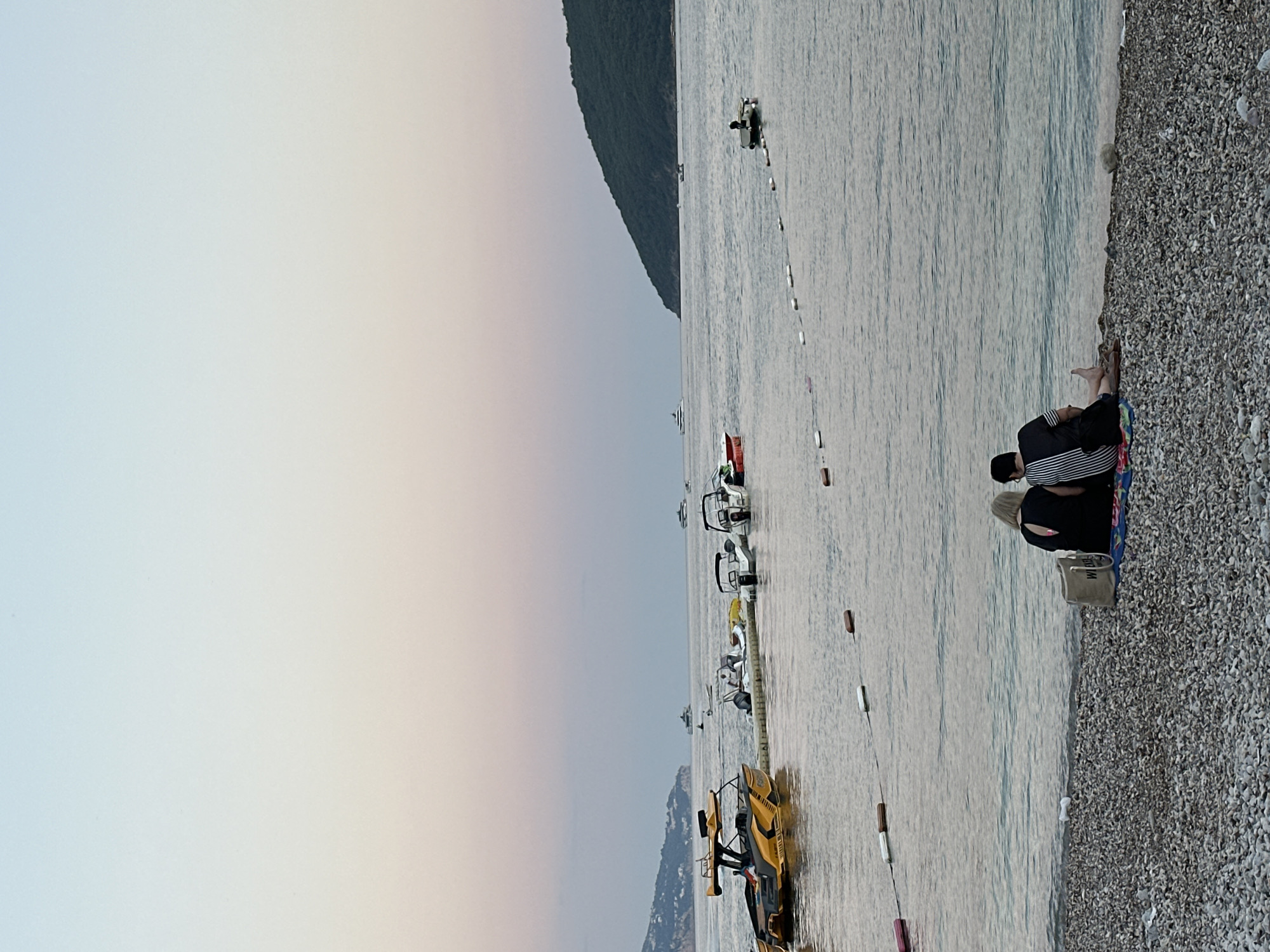 An outdoor, horizontal shot captures a serene evening scene on a pebble beach in Budva, Montenegro. In the foreground, two individuals sit close together, facing the calm sea and the horizon. The person on the left has blonde hair and wears a black top with a cutout on the back. The person on the right has dark hair and wears a black and white striped shirt and dark shorts. A patterned blanket is spread out beneath them, and a beige tote bag rests beside them.
The water is a muted blue-grey, reflecting the soft pastel hues of the sky, which transitions from a pale blue at the top to a soft pink and peach near the horizon. A line of white and brown buoys stretches across the water, marking a boundary.
Further out in the sea, several boats are visible. To the left, a striking yellow and black boat is docked near a floating pontoon. Several other smaller white boats are moored nearby. In the distance, a large green, tree-covered hill dominates the right side of the frame, with a settlement visible on its slopes. On the far horizon, a large white yacht is visible, and other distant boats dot the seascape. A small, dark-colored boat with a single occupant is positioned in the middle ground, to the right of the buoys. The overall atmosphere is tranquil, indicative of a peaceful evening by the Adriatic Sea.