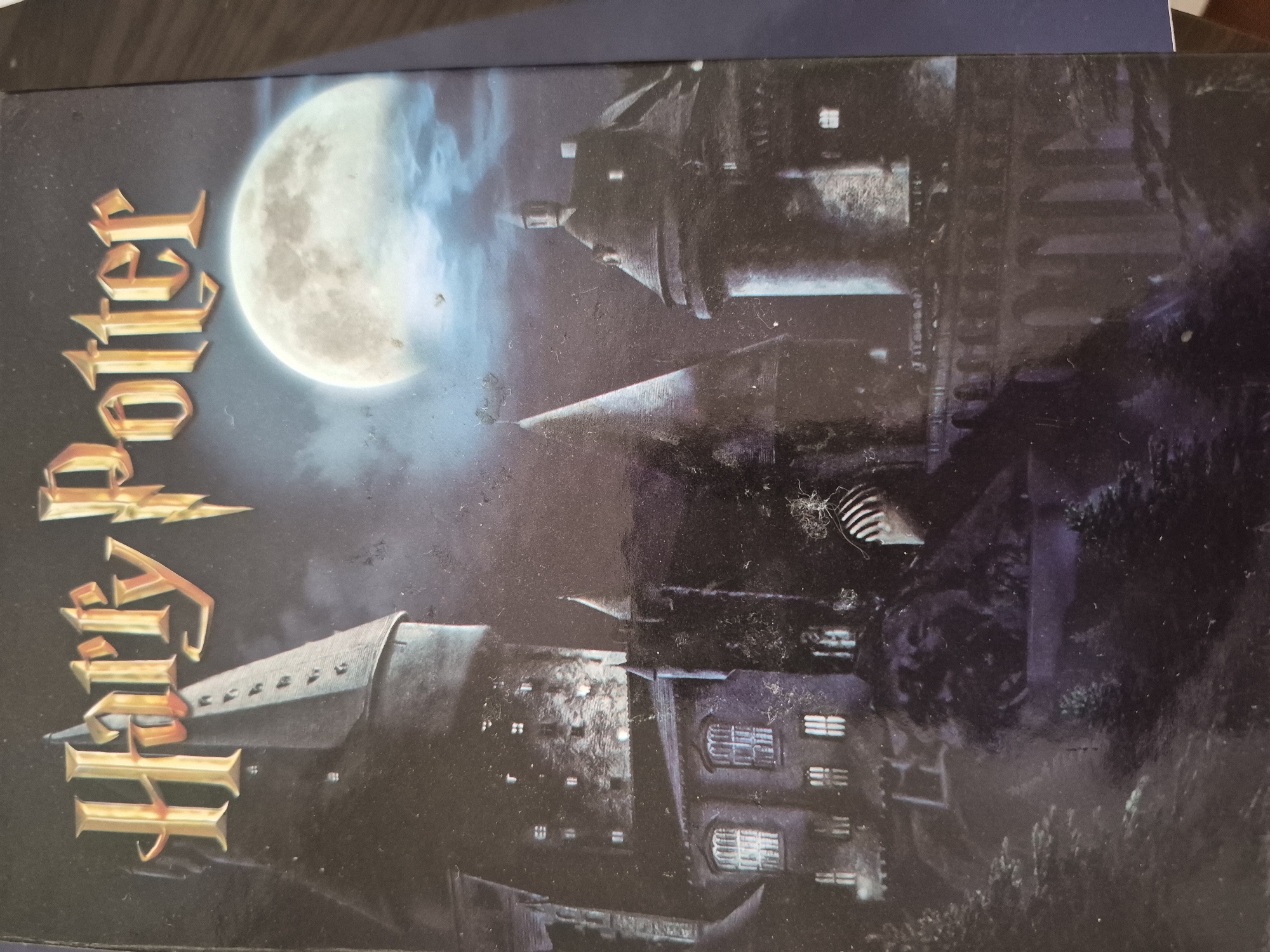 A dark and atmospheric image showcases a grand, multi-turreted castle at night, strongly resembling Hogwarts. A large, luminous full moon dominates the upper left portion of the dark sky, casting a soft glow. Dark clouds or mist swirl around the castle's spires and battlements, enhancing the mystical and somewhat eerie ambiance. The castle features numerous windows, some faintly lit, and intricate architectural details including towers and varied rooflines. Along the left side of the image, the title "Harry Potter" is prominently displayed in a stylized, golden, embossed-like font. The scene is static, depicting a fantastical setting under a night sky. The location context provided (Yerevan, Armenia) refers to the physical location where the photograph was taken, not the fictional scene itself.