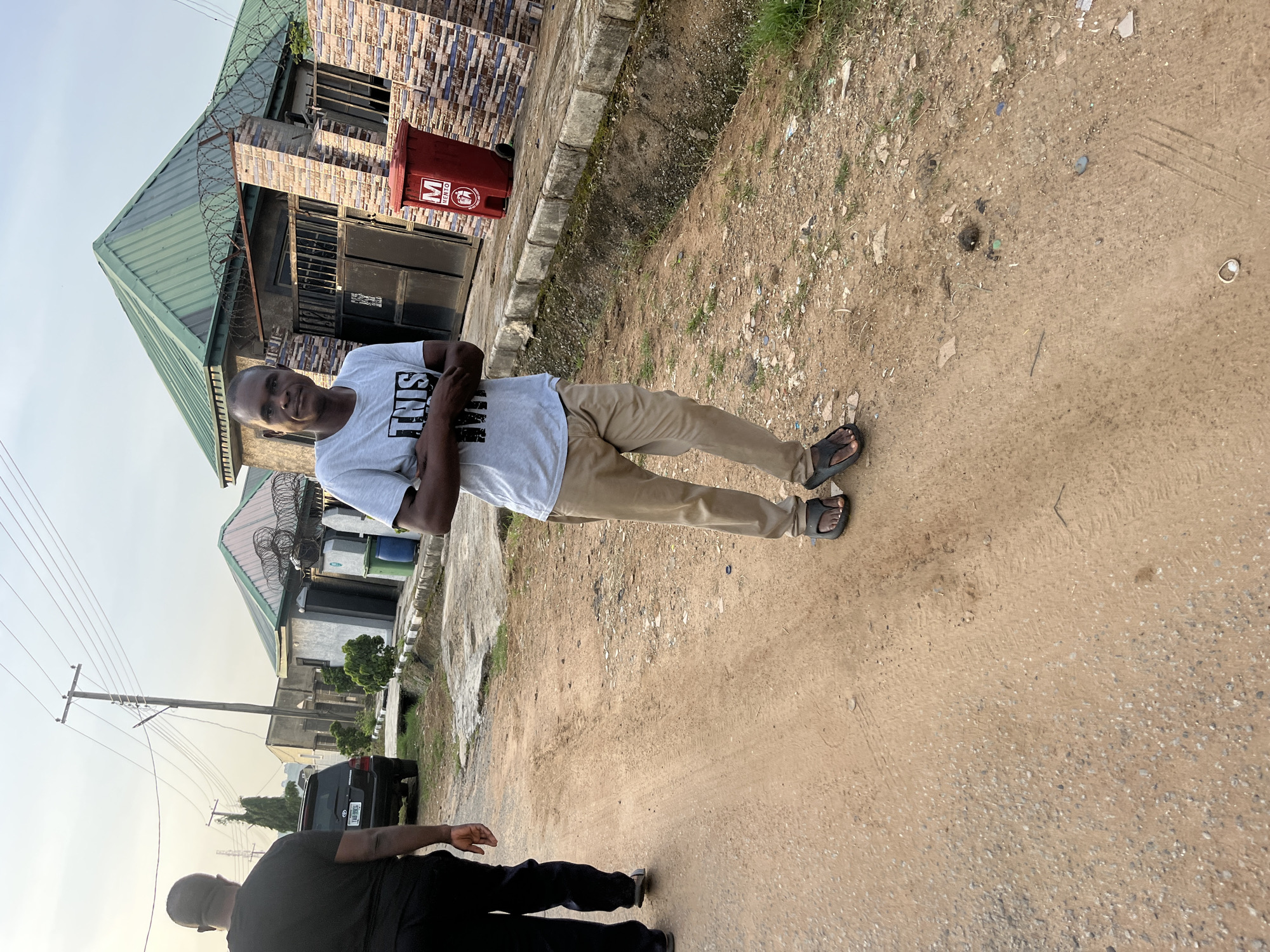 The image shows an outdoor scene in what appears to be a residential area in Abuja, Nigeria. In the foreground, a man is standing on a dirt road, facing the camera. He is wearing a light gray t-shirt with black lettering and khaki pants. His arms are crossed in front of him. To the left of him, another man is walking away from the camera, wearing a black t-shirt and black pants. Behind them, a dark-colored SUV is parked on the side of the road. Several buildings with green roofs and brick accents are visible in the background. A red trash bin with a logo is located near one of the buildings. The overall environment is dusty and somewhat unkempt, consistent with many developing urban areas. The sky is overcast, suggesting it is either early morning or late afternoon.