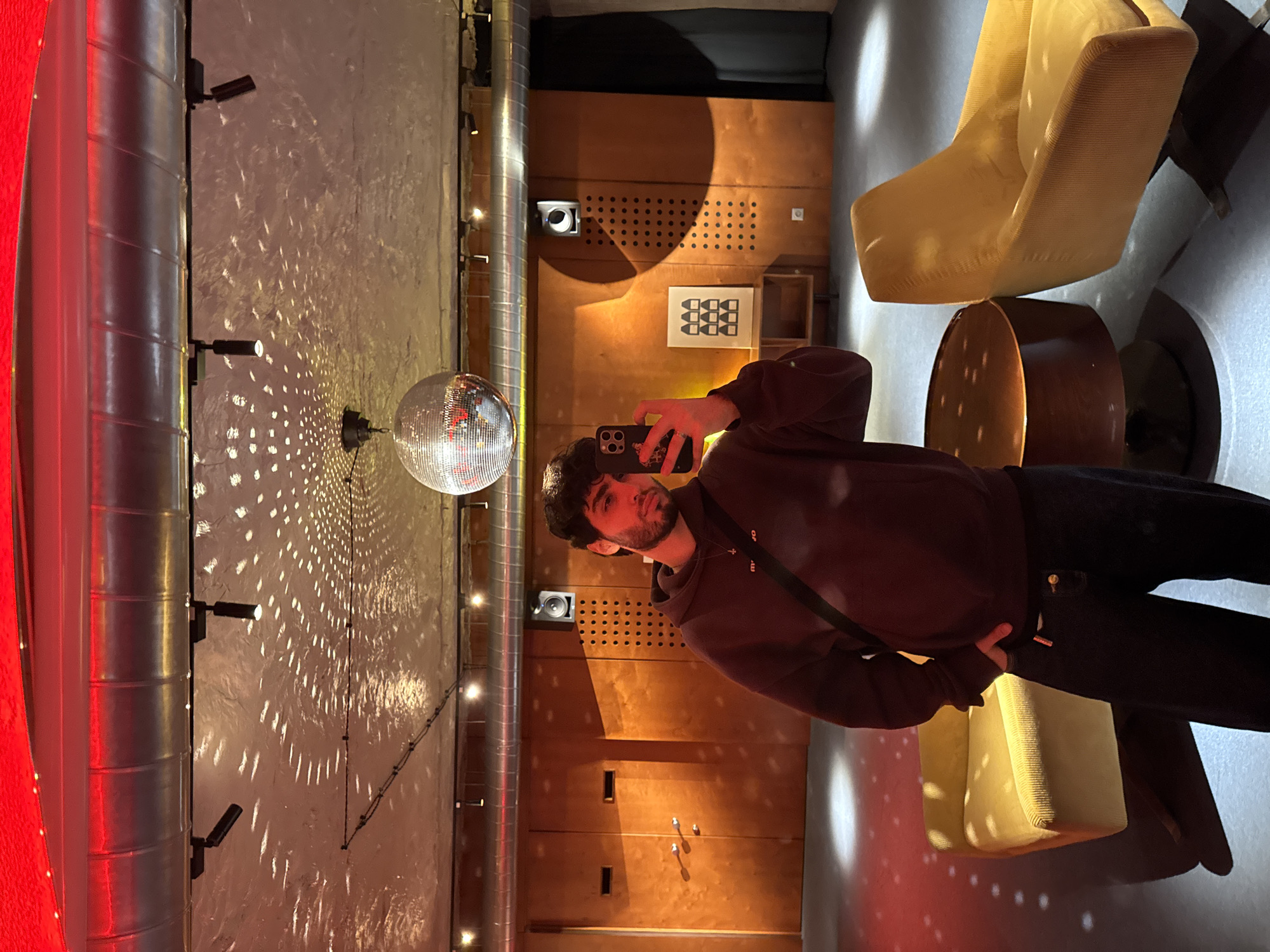 A man stands indoors, possibly at a nightclub or bar in Yerevan, Armenia, holding a smartphone up to take a selfie near a disco ball. The disco ball casts light patterns on a wall behind him. The venue features modern decor with chairs, wooden paneling, and cylindrical ductwork visible. The man is wearing a dark hoodie and pants, along with a crossbody bag.