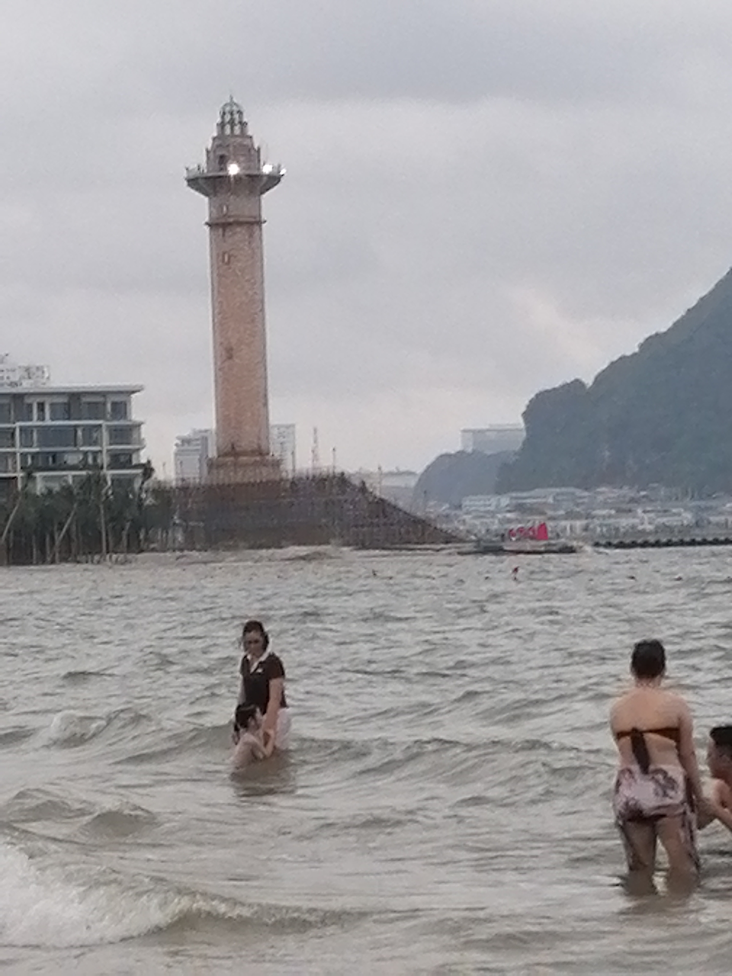 The image depicts a coastal scene with people wading in the sea under an overcast sky. In the foreground, several individuals are partially submerged in the choppy, grey-green water. To the left, a woman wearing a dark top and light bottoms stands, holding a small child. Further to the right, another woman is viewed from behind, wearing a dark bikini top and patterned shorts, standing next to a partially visible man or boy.
In the mid-ground, a tall, light-colored tower, resembling a lighthouse, rises prominently from a large, dark, elevated base. The tower has visible lights near its top section and an ornate spire. To the left of the tower, modern buildings with multiple stories and a white facade are visible, partially obscured by trees. To the right of the tower and extending into the background, a large, dark mountain range dominates the horizon. Below the mountain, numerous structures line the distant shore, and a small red object is seen in the water. The sky is a uniform, light grey, indicating cloudy weather. The overall setting is a populated coastal area, consistent with Bai Chay Ward, Vietnam. No discernible text is visible.