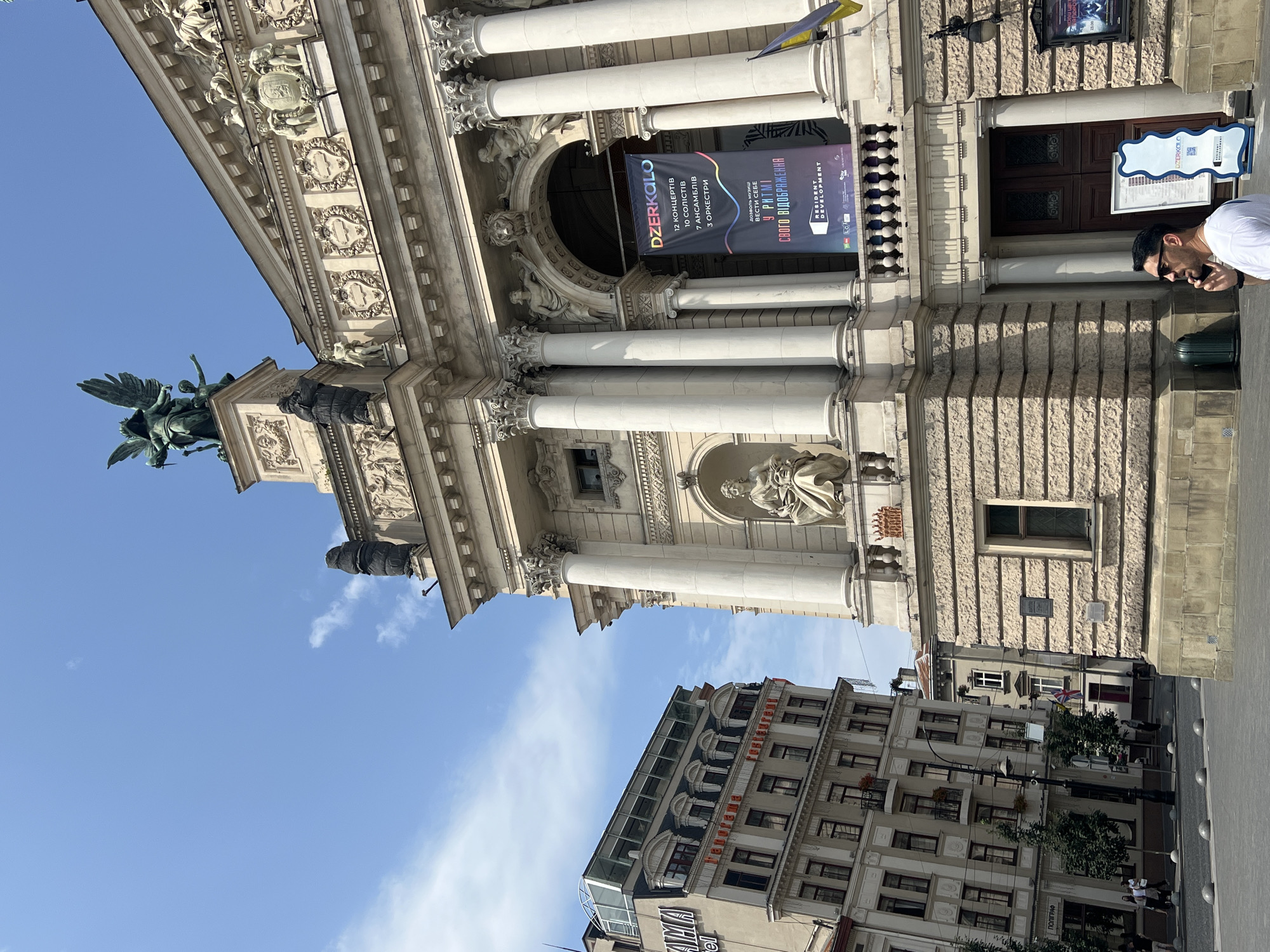The image depicts the Lviv Opera House, a prominent landmark in Lviv, Ukraine, on a sunny day. The grand building features classical architecture with large columns, intricate sculptures, and a prominent banner advertising an event. To the left of the opera house, a modern building with a glass roof stands, contrasting with the historical architecture. A man in a white t-shirt and sunglasses is walking on the street, talking on his phone, with a trash bin and a signpost nearby. The street is paved with cobblestones, and several small, spherical bollards line the sidewalk. The sky is clear blue with a few scattered clouds.