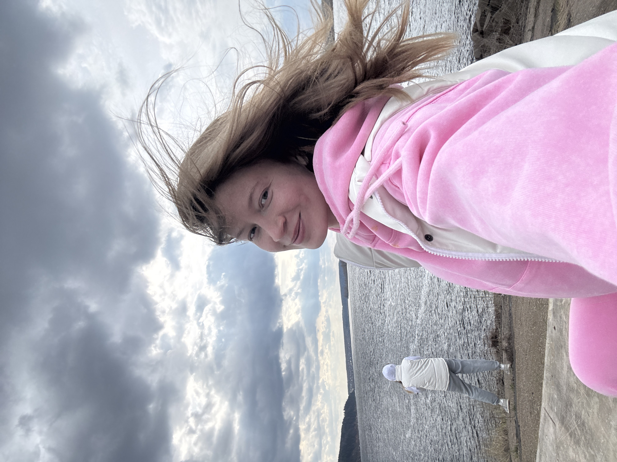 The image captures a dynamic, tilted selfie-style shot of a young person outdoors, with a vast body of water and a cloudy sky in the background. The main subject, a young person with light brown hair, smiles directly at the camera. Their hair is actively blowing upwards and to the left, indicating windy conditions. They are wearing a bright pink, velvet-textured hooded sweatshirt over a white or light-colored jacket.
In the mid-ground, a second person is visible in the distance, standing near the water's edge with their back to the camera. This person is dressed in light grey pants, a white vest, and a white hood or hat.
The setting appears to be along the bank of a large body of water, possibly a reservoir or river, consistent with the location context of Chaikovsky, Russia (Kama Reservoir). A concrete or paved path runs along the water's edge where the second person stands. On the distant horizon, a faint line of land or hills is visible under the expansive sky.
The sky is largely overcast with dense grey clouds, although patches of brighter white clouds suggest the sun is either breaking through or diffused behind them. The overall lighting is soft and natural, typical of a cloudy day. The image has a casual and joyful feel, conveyed by the main subject's smile and the lively movement of their hair. No visible text is present in the image.