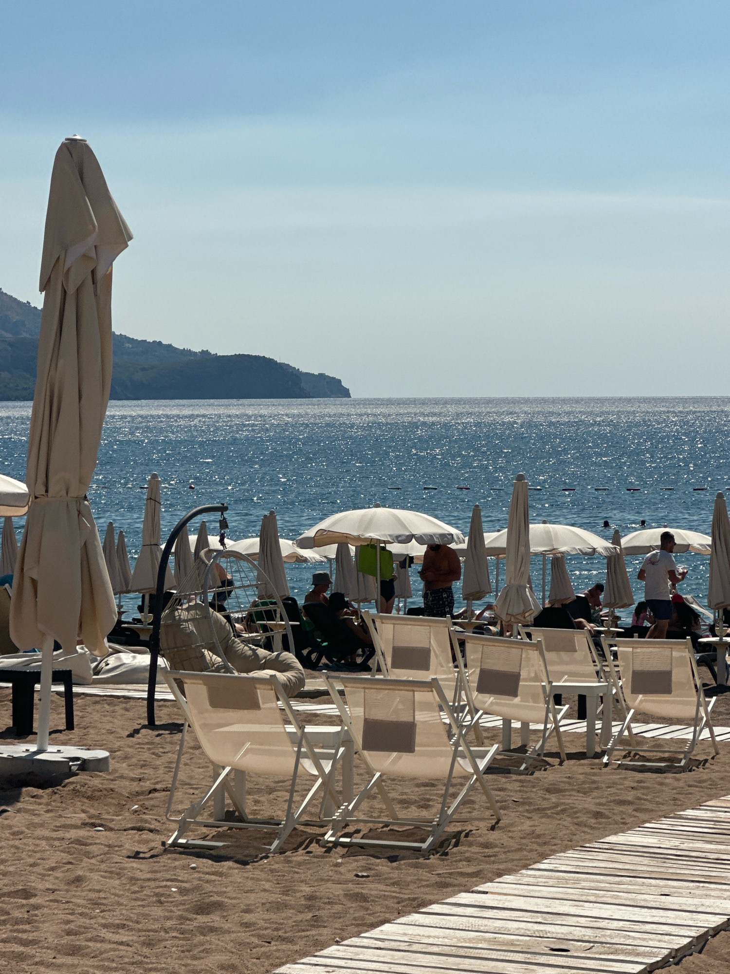 A sunny day at the beach in Boreti, Montenegro features a shoreline with sandy ground and a wooden walkway. Numerous closed and open white umbrellas are set up on the beach, with rows of empty white lounge chairs in the foreground. Beyond the chairs, several people are visible, some sitting under umbrellas, while others are standing or walking. The sea is calm and blue, with sunlight glinting off the surface. In the distance, a tree-covered hill slopes down to the water. The sky is clear and pale blue.