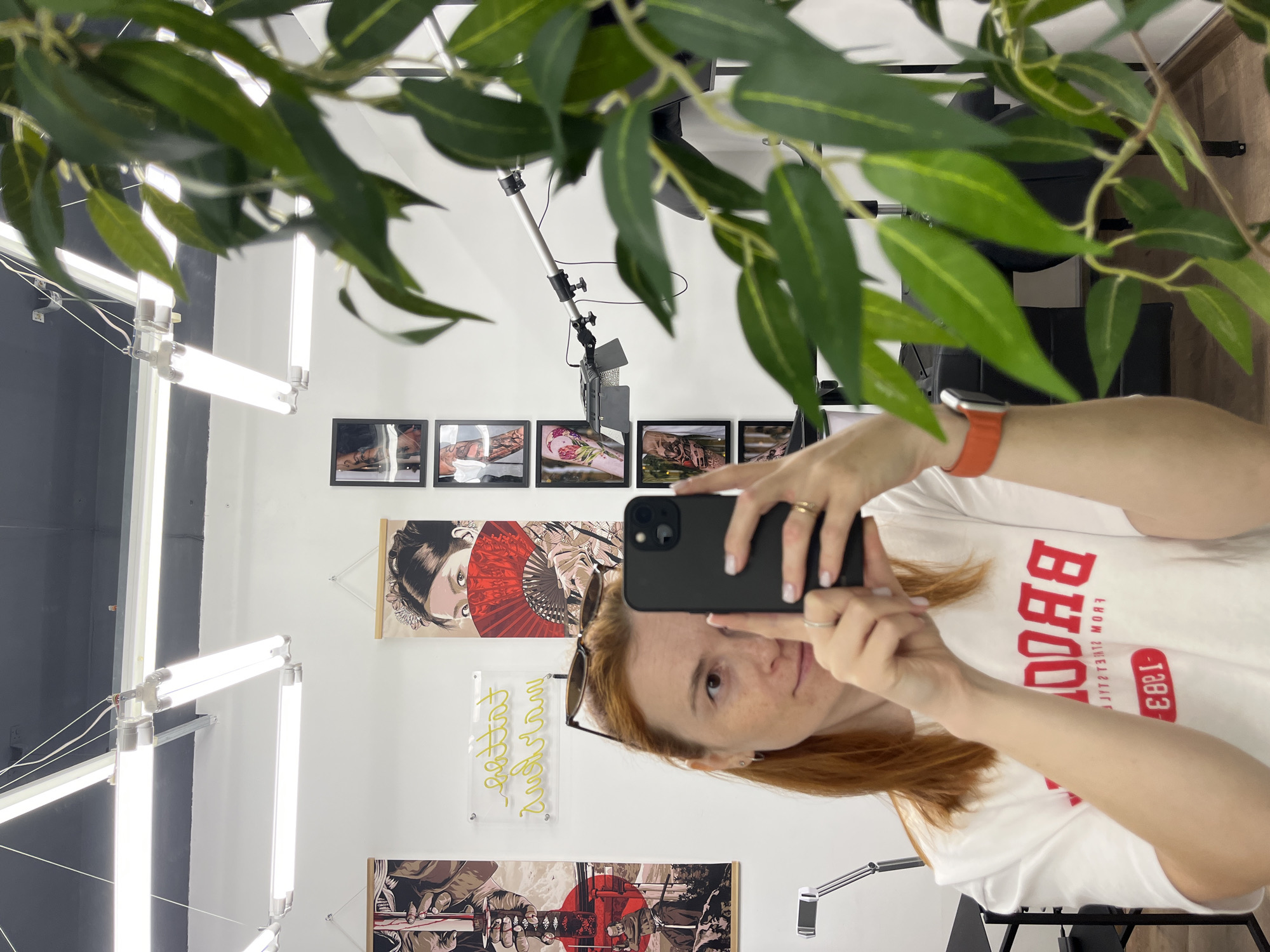 A young Caucasian woman with reddish-brown hair, wearing a white t-shirt with red lettering that reads "BROOKLYN", takes a selfie in a mirror. She is wearing sunglasses pushed up on her head, a silver ring on her left ring finger, and an orange smartwatch. The background appears to be a tattoo parlor. The wall features several framed tattoo art prints, a neon sign with cursive lettering, and two large hanging posters. Overhead, fluorescent lights illuminate the space. A large potted plant with green leaves partially obstructs the view on the right side of the frame. The woman is holding a black smartphone to take the picture. The overall impression is a casual, modern aesthetic within a professional setting. This image is not related to the city of Chaikovsky, Russia.