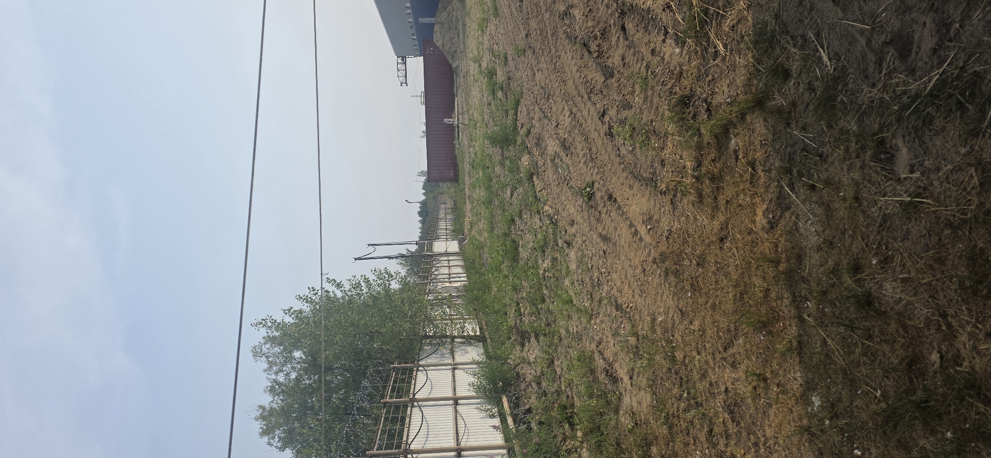 The image shows an outdoor scene with a fence along the left side and a building in the background on the right. The fence is made of corrugated metal panels with vertical posts and barbed wire on top. Trees are visible behind the fence. In the middle ground, there is a large red shipping container. The ground in the foreground and middle ground appears to be unpaved and covered with sparse vegetation and tire tracks. The sky is a light blue with a few wispy clouds. The scene appears to be in an industrial or undeveloped area. There is no visible indication of the specific location within the city of Nizhnevartovsk, Russia.