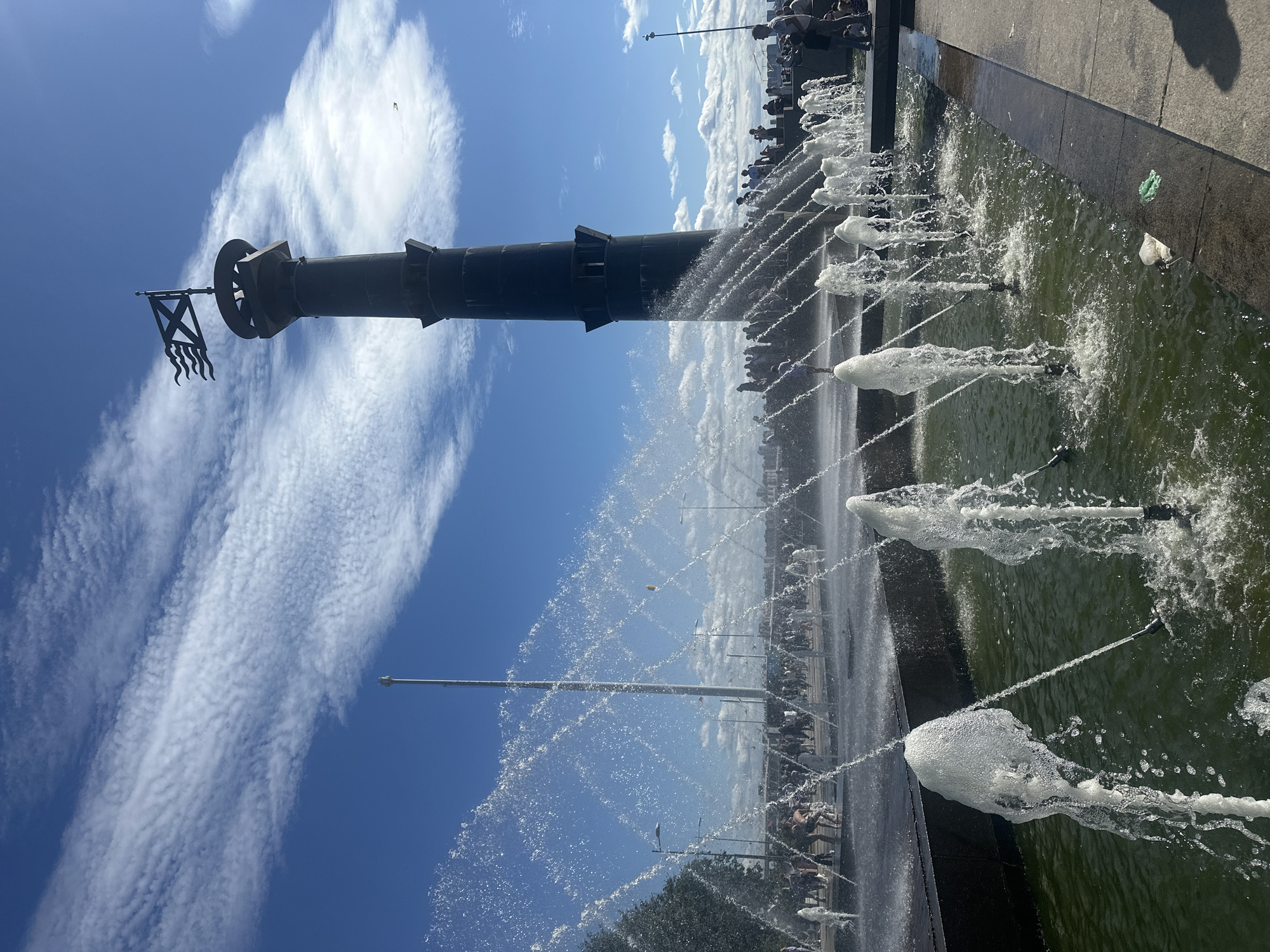 The image depicts a sunny day at a waterfront park in Saint Petersburg, Russia. In the foreground, a large public fountain sprays water high into the air, creating a dynamic visual effect. The water is a green hue, suggesting it is a large, possibly decorative, pool. In the midground, a diverse crowd of people are gathered, many of them dressed casually, enjoying the outdoor space. Some appear to be walking, while others are standing and observing the surroundings. Behind the crowd, a tall, dark, cylindrical monument stands prominently. It is topped with a flag featuring a diagonal cross and wavy lines, likely a historical or naval ensign. The sky is a vibrant blue, streaked with wispy white clouds, indicating good weather. The overall scene suggests a lively public gathering in an urban setting, likely a popular tourist or recreational area.