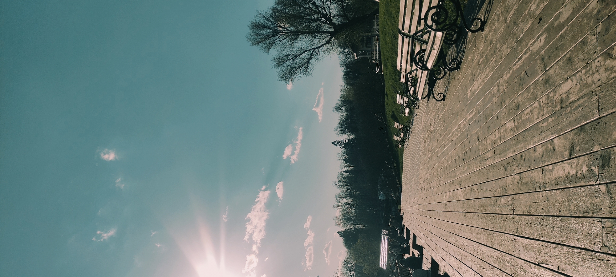 The image shows an outdoor scene in Grebnevo, Russia. A weathered wooden walkway extends into the distance, lined with park benches. The sky is light blue with sparse white clouds, and the sun is shining brightly. Trees and greenery border the walkway.