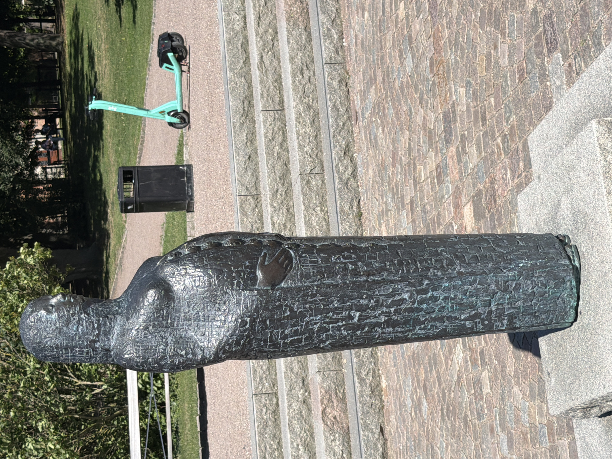 In Lund, Sweden, a weathered bronze statue of a pregnant woman stands on a stone pedestal in a park. The statue is depicted in a long, textured robe, with one hand resting on her abdomen. Behind the statue, a black trash can sits next to a bright green electric scooter. In the background, there are stone steps leading up to a grassy area with trees and benches where two people are seated. The ground is paved with cobblestones.