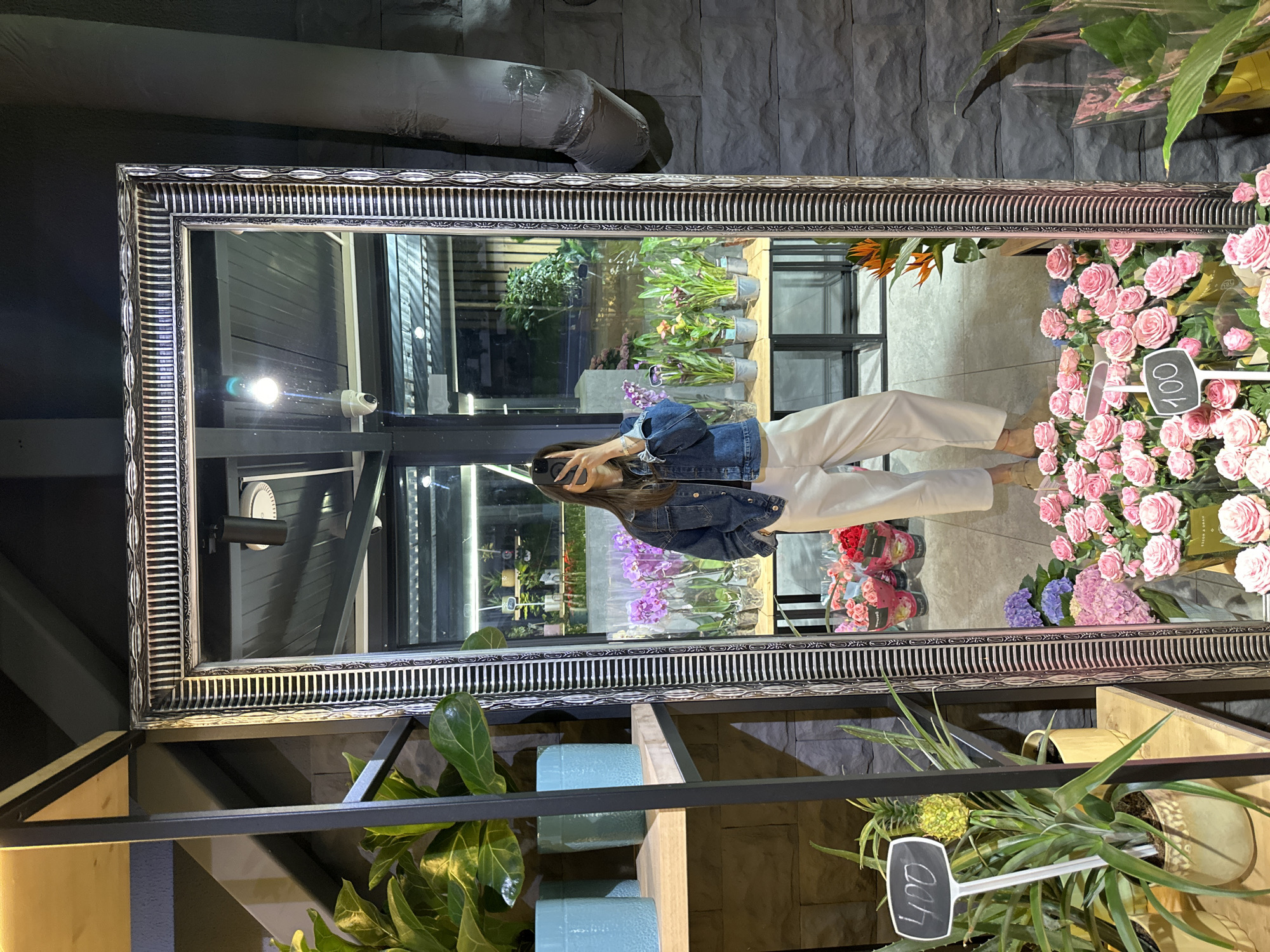 A person stands in front of a large ornate mirror, taking a selfie. They are wearing a denim jacket and white pants. The mirror reflects a flower shop, with various plants and floral arrangements visible. In the foreground of the reflection, there are bouquets of pink and red roses, as well as purple hydrangeas. Behind them, shelves hold potted plants. A sign with the number "100" is placed in front of the roses. To the left of the mirror, in the real environment, are shelves with blue pots and green plants. The wall behind the mirror is made of dark stone. This scene is located in the city of Chișinău, Moldova.