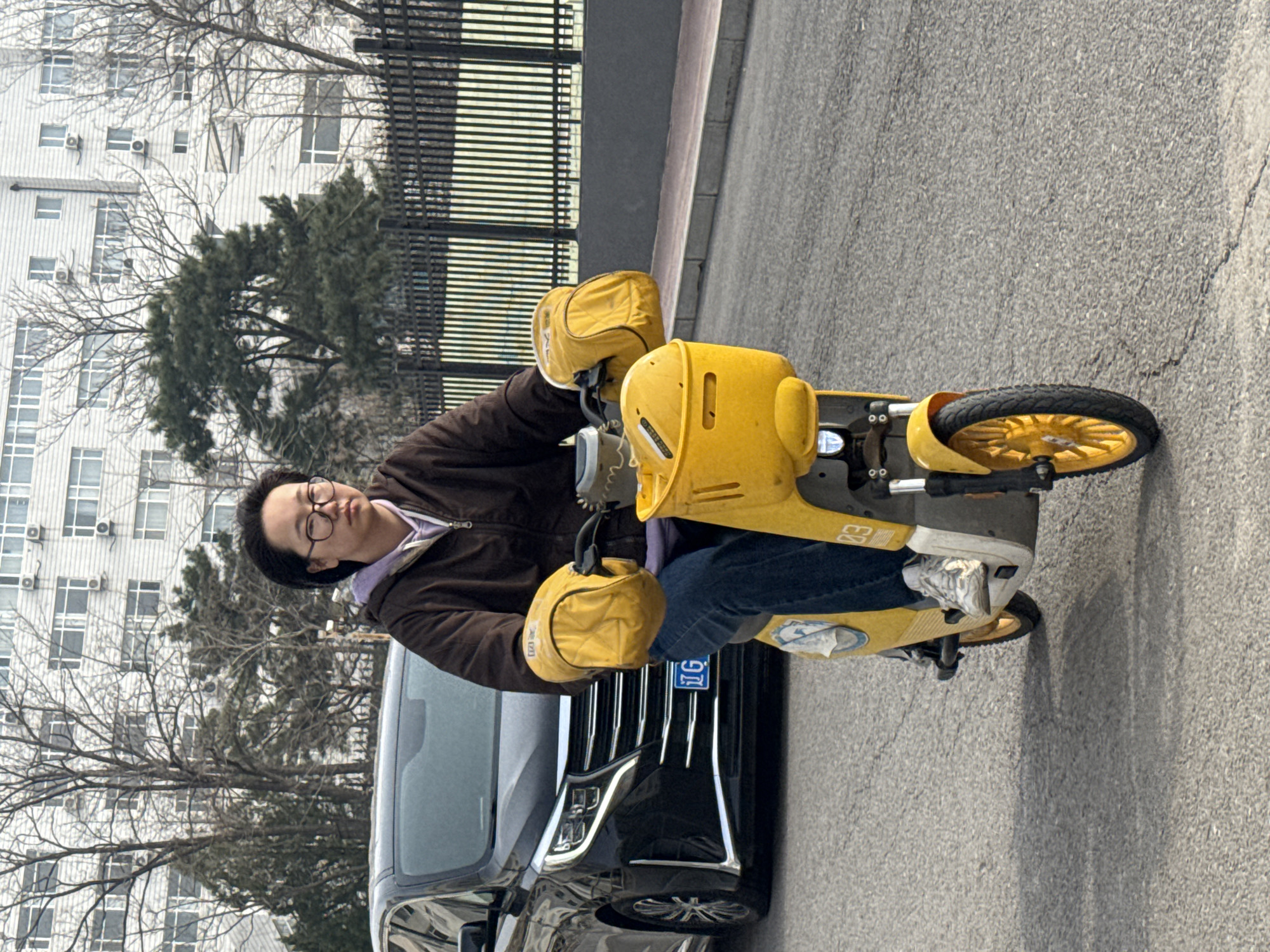 A woman wearing glasses, a brown jacket over a purple hoodie, and blue jeans is riding a bright yellow electric scooter with yellow hand covers. She is parked next to a black car with a license plate. In the background is a building with many windows and air conditioning units, trees, and a black fence. The photo appears to be taken on a street in Zhongtun, China.