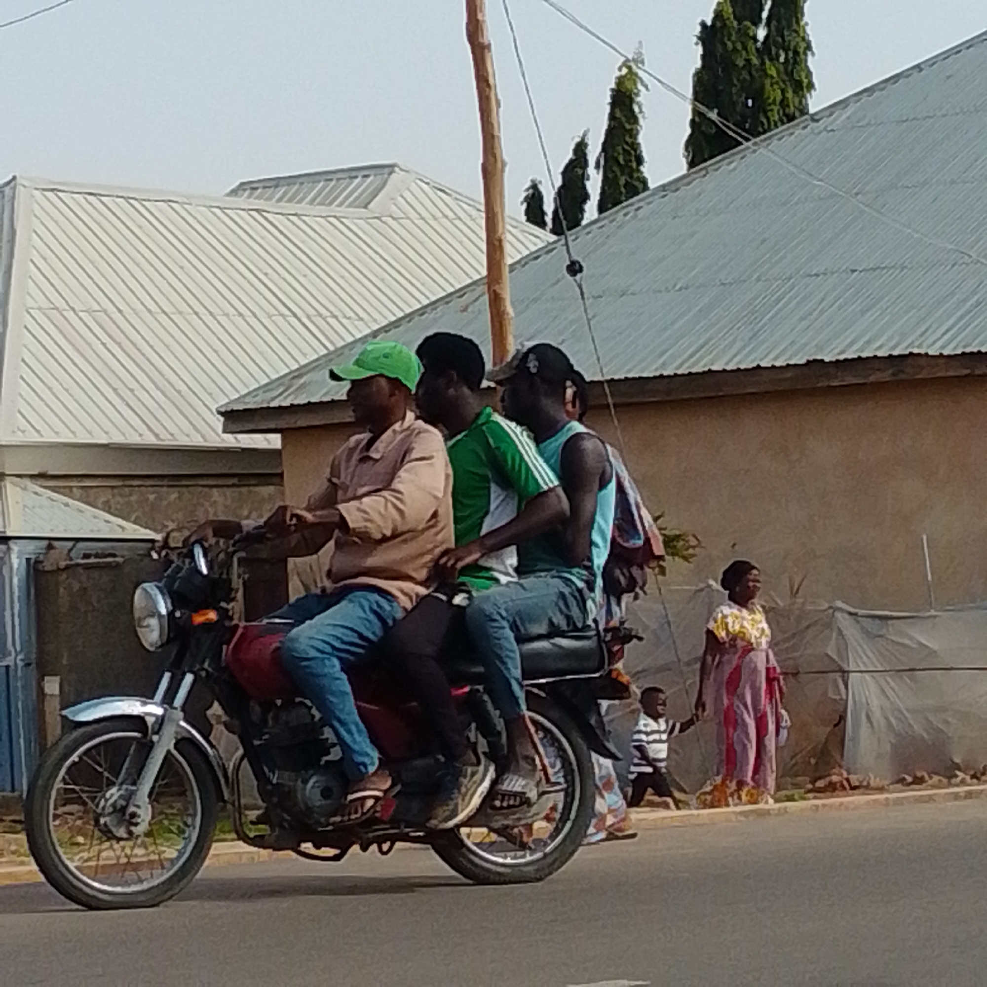The image depicts a street scene in Gombe, Nigeria. In the foreground, three men are riding on a motorcycle. The rider in front, wearing a light brown shirt and blue jeans, has a green cap. The middle passenger is wearing a green and white striped shirt, and the rear passenger is in a blue tank top and jeans. They are all looking forward. Behind them, a woman in a colorful dress and a child in a striped shirt are standing near a white tarp. The background shows buildings with corrugated metal roofs and some trees. The road is paved, and the overall impression is of a typical day in an urban area.