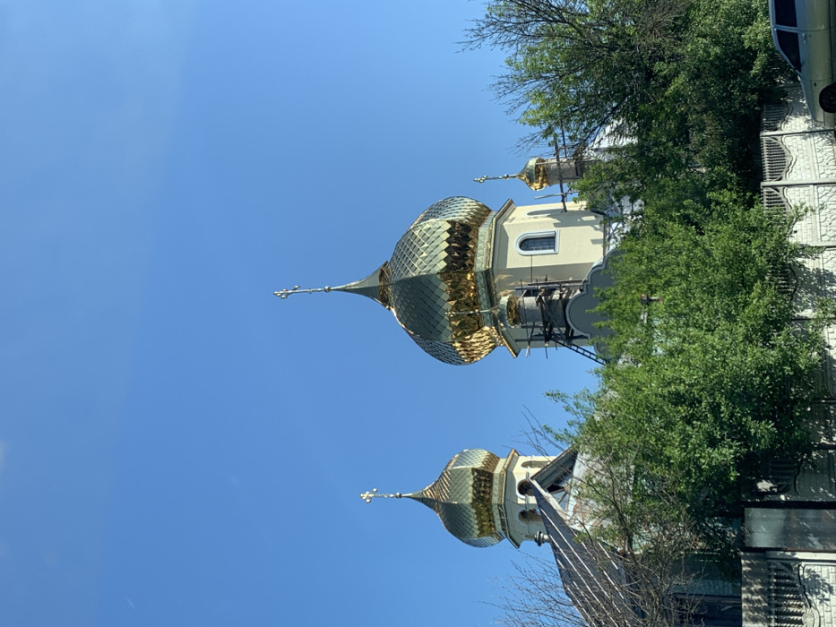 The image shows the front of a church with three gold domes in Kolomak, Ukraine, located at the coordinates 49.837 latitude and 35.294 longitude. The church is made of yellow brick with white trim and has a scaffolding structure on its front. It is partially obscured by a thick line of deciduous trees with green leaves. There is a low brick wall with a metal fence in front of the church, and the top of a car is visible in the bottom right corner of the image. The sky is a bright blue with a few scattered white clouds. The only human subjects are those who built the church and scaffolding, who are not visible in the image.