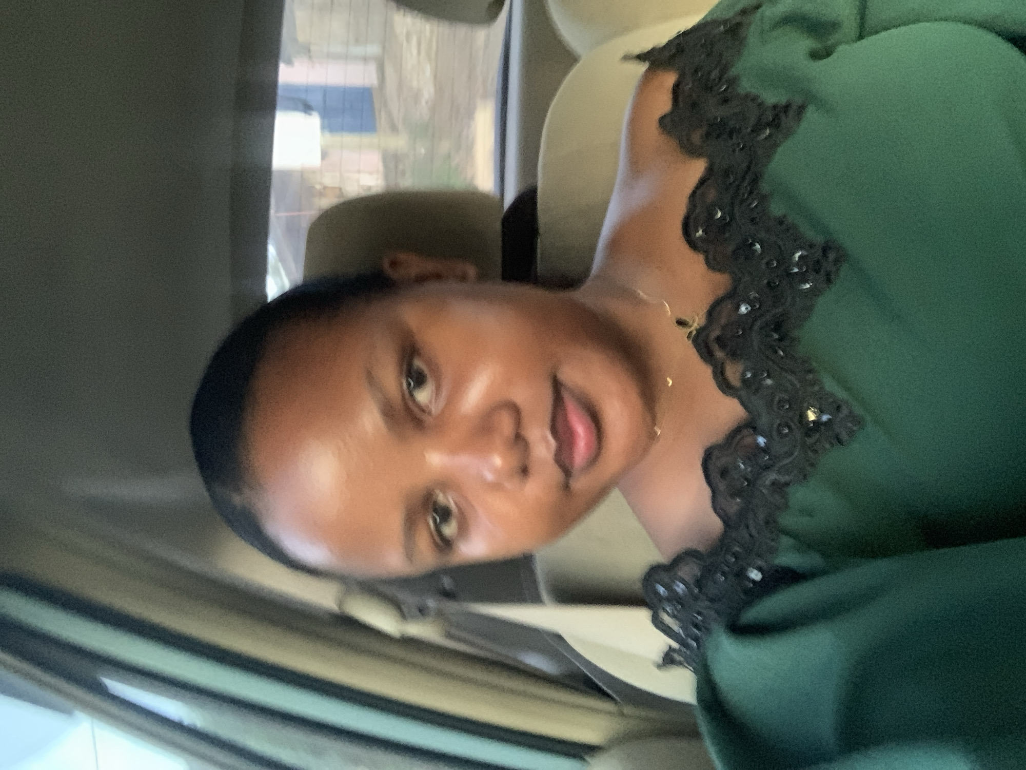 A woman is sitting in a car. She is wearing a green dress with black lace around the neckline and a gold necklace. She is looking at the camera with a neutral expression. The background shows a cityscape through the car window. Given that she is in the city of Dar es-Salaam, Tanzania, the buildings are likely residential or commercial structures typical of the city.