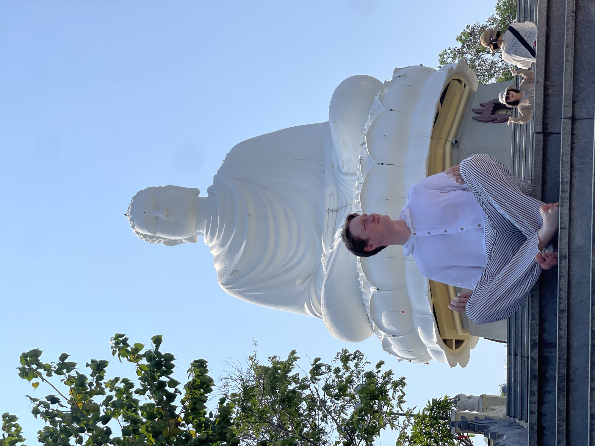 This image, taken in Tây Nha Trang, Vietnam, captures a serene scene centered around a large, white statue of a sitting Buddha. The monumental statue, likely depicting Amitabha Buddha given its meditative posture and hand gesture, rests on a multi-tiered base adorned with lotus petal-like structures and subtle golden accents, set against a clear, bright blue sky.
In the foreground, a person is seated on steps, facing the Buddha statue. They are dressed in a loose white long-sleeved shirt and light-colored striped pants, with their legs crossed and bare feet visible. Their head is tilted back slightly, and their eyes are closed, conveying a sense of meditation, contemplation, or peaceful reverence.
Further up the stairs, two other individuals are partially visible behind the meditating person and the statue's base. One wears a straw hat and a light-colored top, while the other, also in a hat, appears to be pointing or gesturing. Green tree foliage and branches frame the lower and left portions of the image.
The bright sunlight and cloudless sky suggest a clear day, likely morning or late afternoon, contributing to the tranquil atmosphere. The overall scene evokes a sense of spiritual peace and devotion at a significant Buddhist temple or site in Vietnam. There is no visible text in the image.