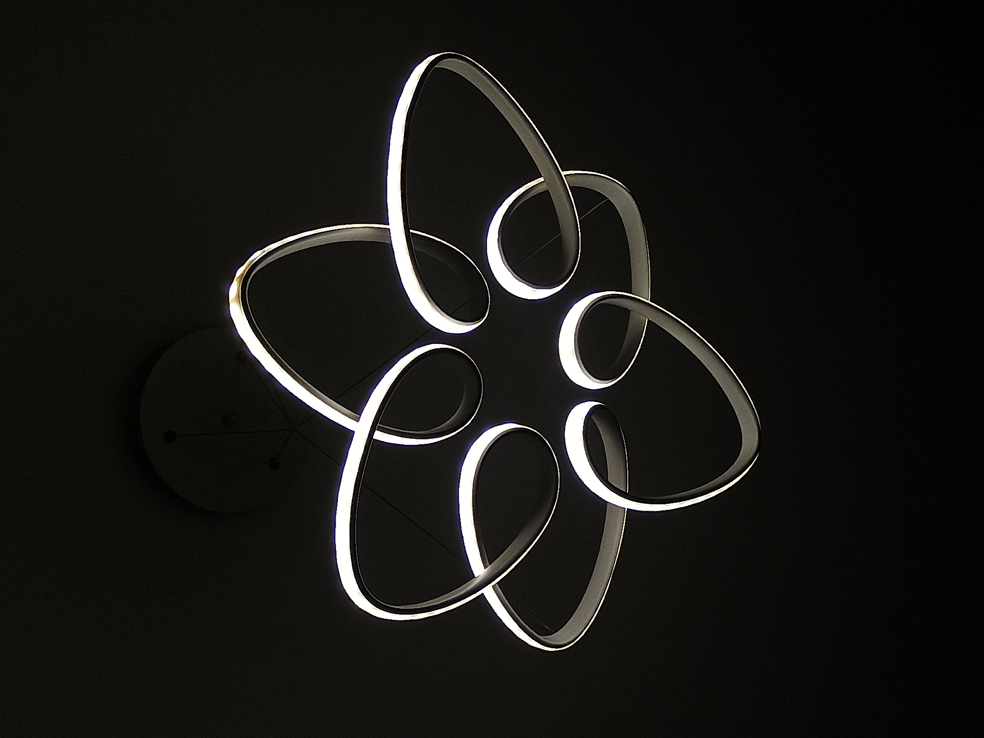 The image shows a modern chandelier against a dark background. The chandelier has a white light source along the inner edge of a curvilinear, abstract, floral-like design. The fixture is circular, with a mounting plate affixed to the ceiling. Fine wires extend from the mounting plate, connecting to the illuminated structure. The lighting fixture is suspended from a ceiling with a dark, plain background. The location in Plovdiv, Bulgaria is not discernible from this image.