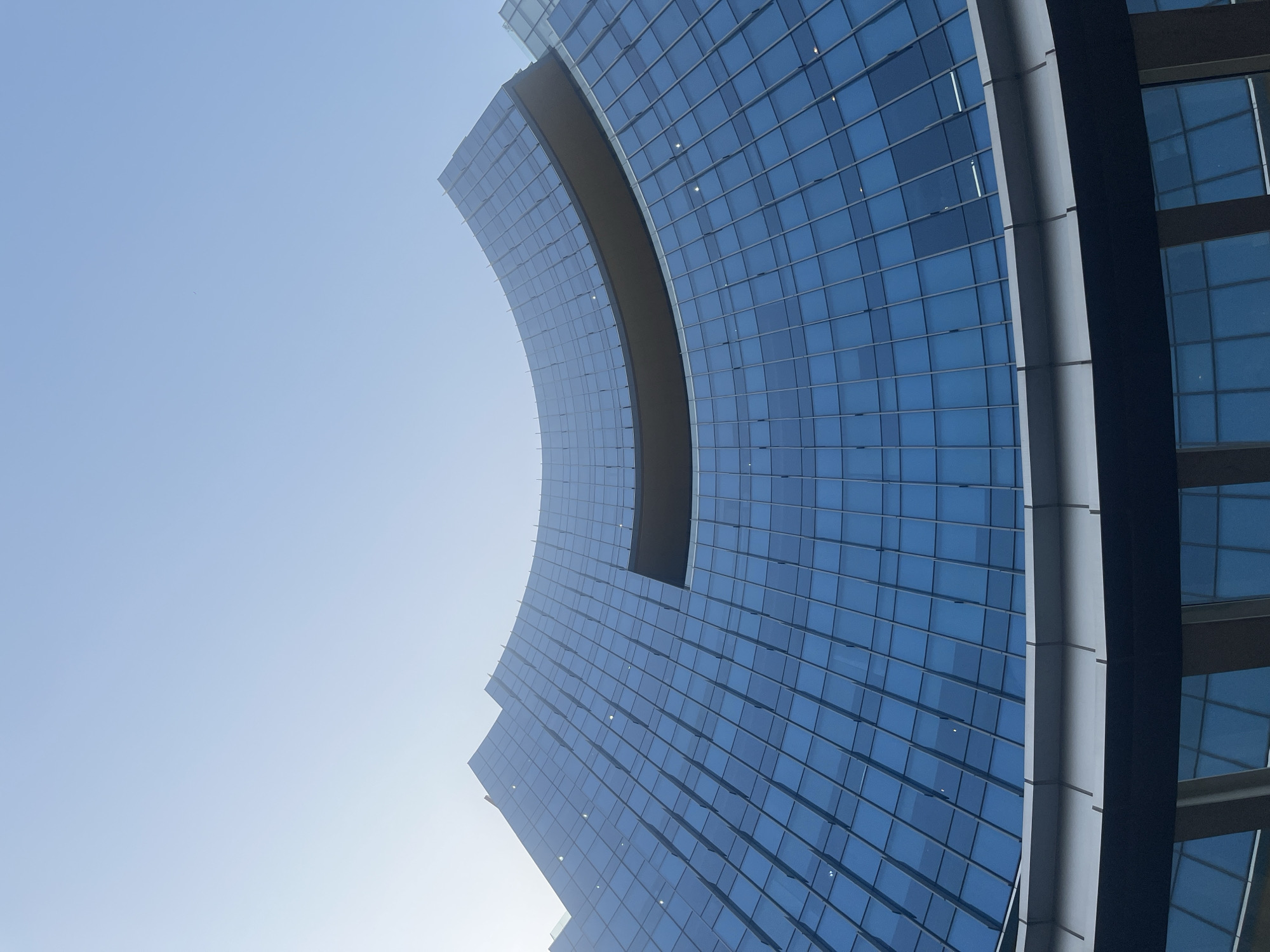 This is a low-angle, upward shot of a modern, curved glass skyscraper in West Jakarta, Indonesia. The building's facade is composed of numerous blue-tinted glass panels, reflecting the clear, pale blue sky above. A dark, curved gap is visible in the upper section of the building, creating a distinctive architectural feature. The lower part of the image shows a portion of the building's base, with darker glass panels and structural supports. No people are visible in the image. The overall impression is one of contemporary architecture against a bright, somewhat hazy sky.