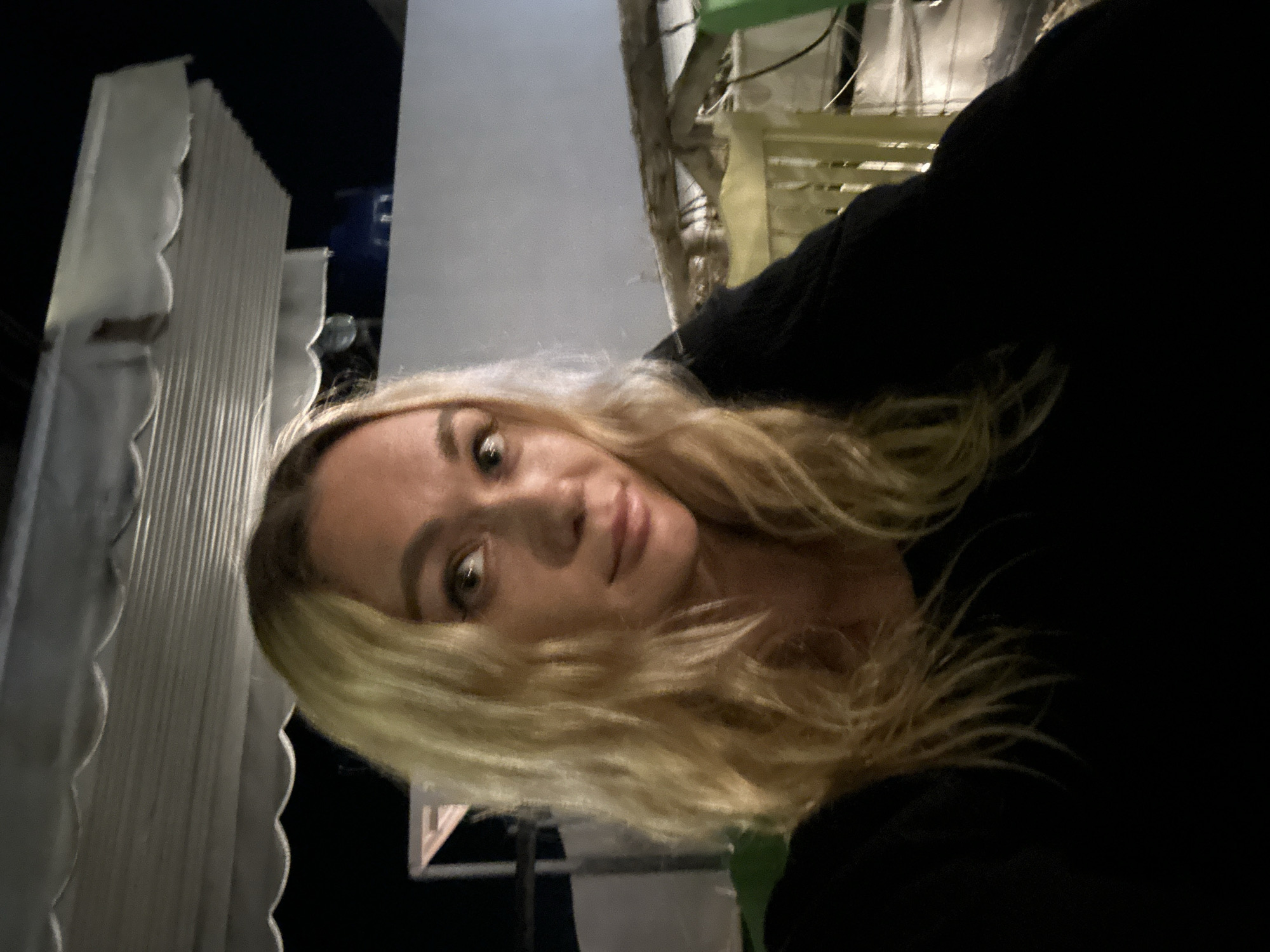 A blonde woman with wavy hair is taking a selfie outdoors at night. She is wearing a black top. In the background, there is a white wall, some awnings, and a yellow chair. The location is not specified in Phu Thuy Ward, Vietnam, nor are there any specific identifiable landmarks.