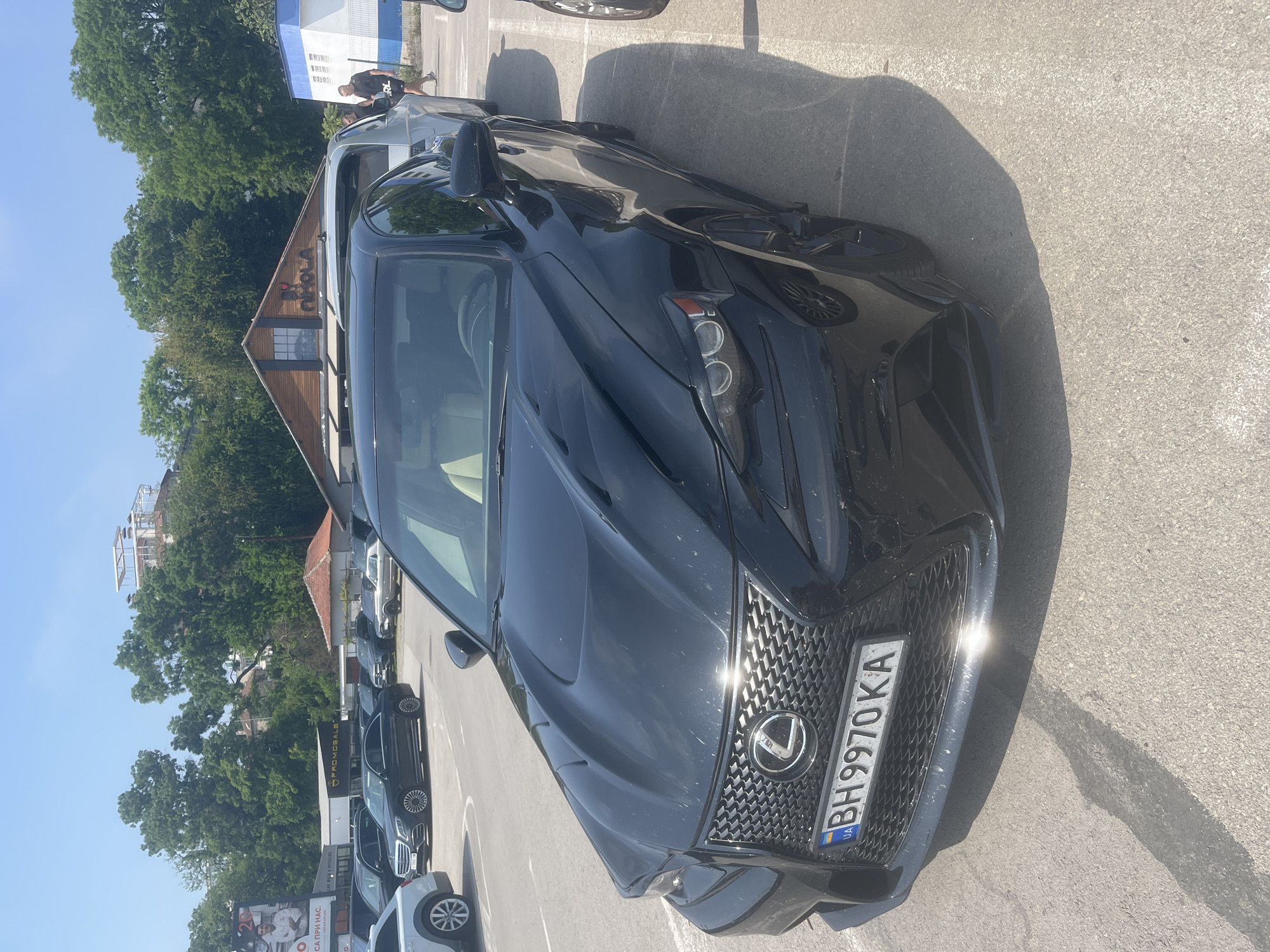 The image displays a black Lexus sedan, likely an IS model, parked outdoors on an asphalt lot under a bright, clear sky. The car is seen from a front-right angle, highlighting its prominent spindle grille with the Lexus emblem and distinctive "L-shaped" daytime running lights. Sunlight creates strong reflections on the car's glossy black paint and the chrome elements of the grille.
The visible license plate on the front of the Lexus reads "BH 9970 KA," featuring a Ukrainian flag and "UA" country code on the left side, indicating Ukrainian registration.
In the background, a distinctive wooden building with a peaked roof is visible, adorned with a sign that clearly spells "OLA." Tall, green deciduous trees frame this building and extend across the mid-ground. To the far left, another building with blue and white sections is partially visible. Further back and to the right of the Lexus, a person wearing dark clothing is walking away from the camera. The parking lot contains several other vehicles, including a light-colored SUV in the foreground and multiple dark sedans parked further back. A large billboard with Cyrillic text, including the phrase "CA ПРИ НАС," can be seen on the left side of the scene.
The scene depicts a sunny day, likely mid-day or early afternoon, given the sharp shadows cast on the asphalt. The setting appears to be a commercial area or a dealership, potentially in Varna, Bulgaria, considering the provided location context, despite the car's Ukrainian license plate.