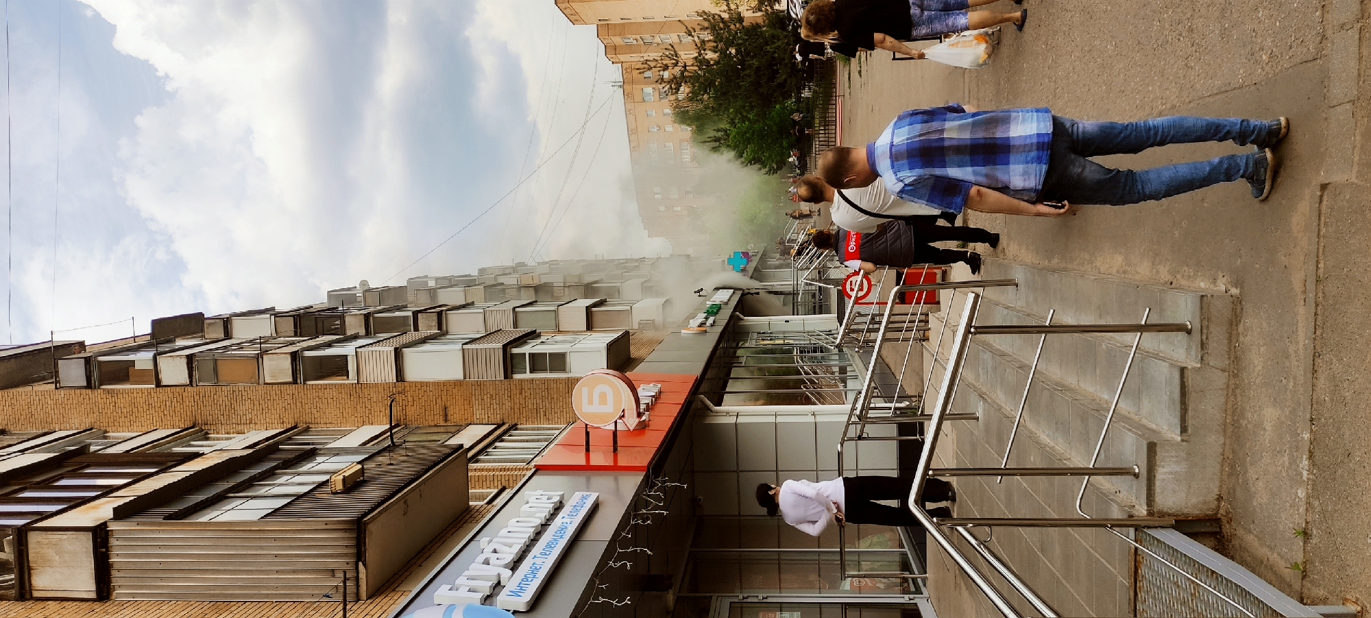 The image captures an outdoor scene on what appears to be a city street, with the location likely being Unknown, Unknown, based on the Cyrillic text on the storefront signs. The foreground features a staircase with metal handrails leading up to a commercial building. This building appears to house multiple businesses, judging by the different signs visible, including what appears to be a television and internet service provider.
Several people are present in the image. Some are walking on the sidewalk, their faces obscured. One man is wearing a blue plaid shirt and jeans. There are individuals standing near the commercial building observing something out of frame.
The environment shows signs of an ongoing incident, likely a fire, as thick smoke billows out from the top of a nearby multi-story building. The sky is overcast with patches of blue visible. The building architecture is characterized by repetitive, modular units or balconies.
The interaction between the people and the environment is one of observation and reaction to the ongoing fire event. Some individuals are stopping to watch, while others continue walking, seemingly unfazed.