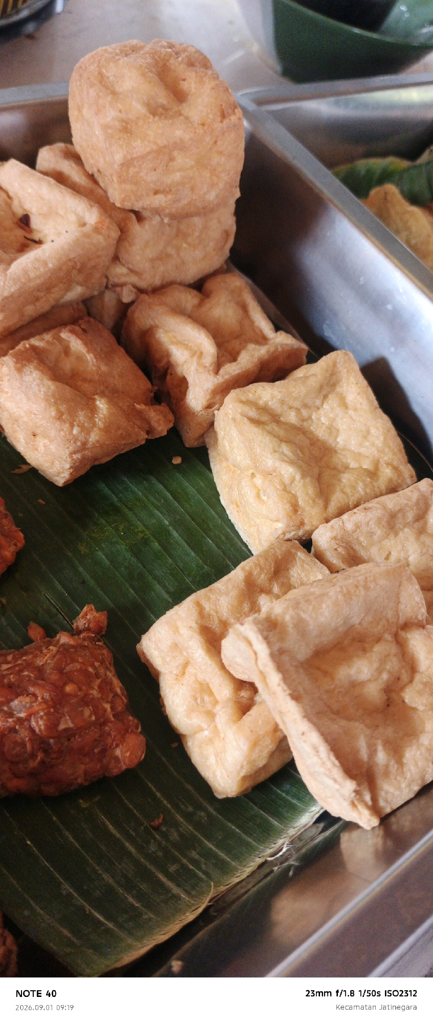 A close-up, eye-level shot shows a selection of fried tofu and tempeh arranged in a metal tray. The food is predominantly golden brown, with some pieces appearing more deeply fried than others. The tofu is cut into cubes and irregular shapes, some stacked upon each other. The tempeh is a darker, reddish-brown, and appears more textured. The food is resting on a bed of large, green banana leaves, which are slightly wilted. The background is blurred, showing the edge of the metal tray and hints of other dishes or containers, suggesting a food stall or market setting. The bottom of the image contains text indicating the camera model ("NOTE 40"), the date and time ("2026.09.01 09:19"), and the camera settings ("23mm f/1.8 1/50s ISO2312"). The text also specifies the location as "Kecamatan Jatinegara," which is a subdistrict in East Jakarta, Indonesia.
