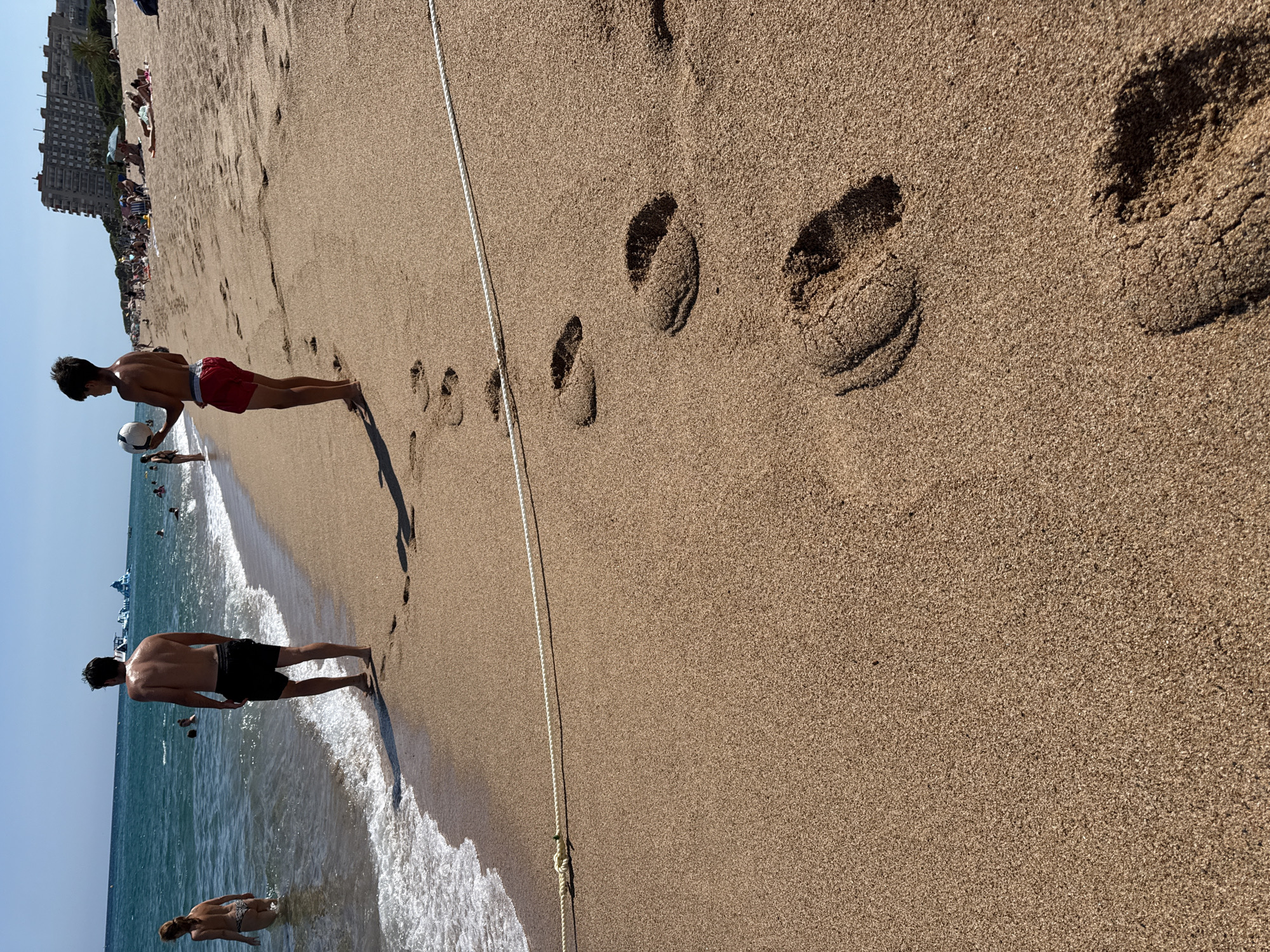 The image depicts a beach scene in Blanes, Spain. In the foreground, distinct footprints are imprinted in the golden sand. A white rope lies across the sand, marking a boundary. Two individuals are visible near the water's edge: a person in black shorts facing away from the camera, and a younger individual in red shorts holding a white soccer ball, also facing away. The sea, with gentle waves, stretches towards the horizon. In the background, other beachgoers, umbrellas, and buildings are scattered along the shoreline, under a clear blue sky. The overall impression is a sunny day at a popular coastal location.