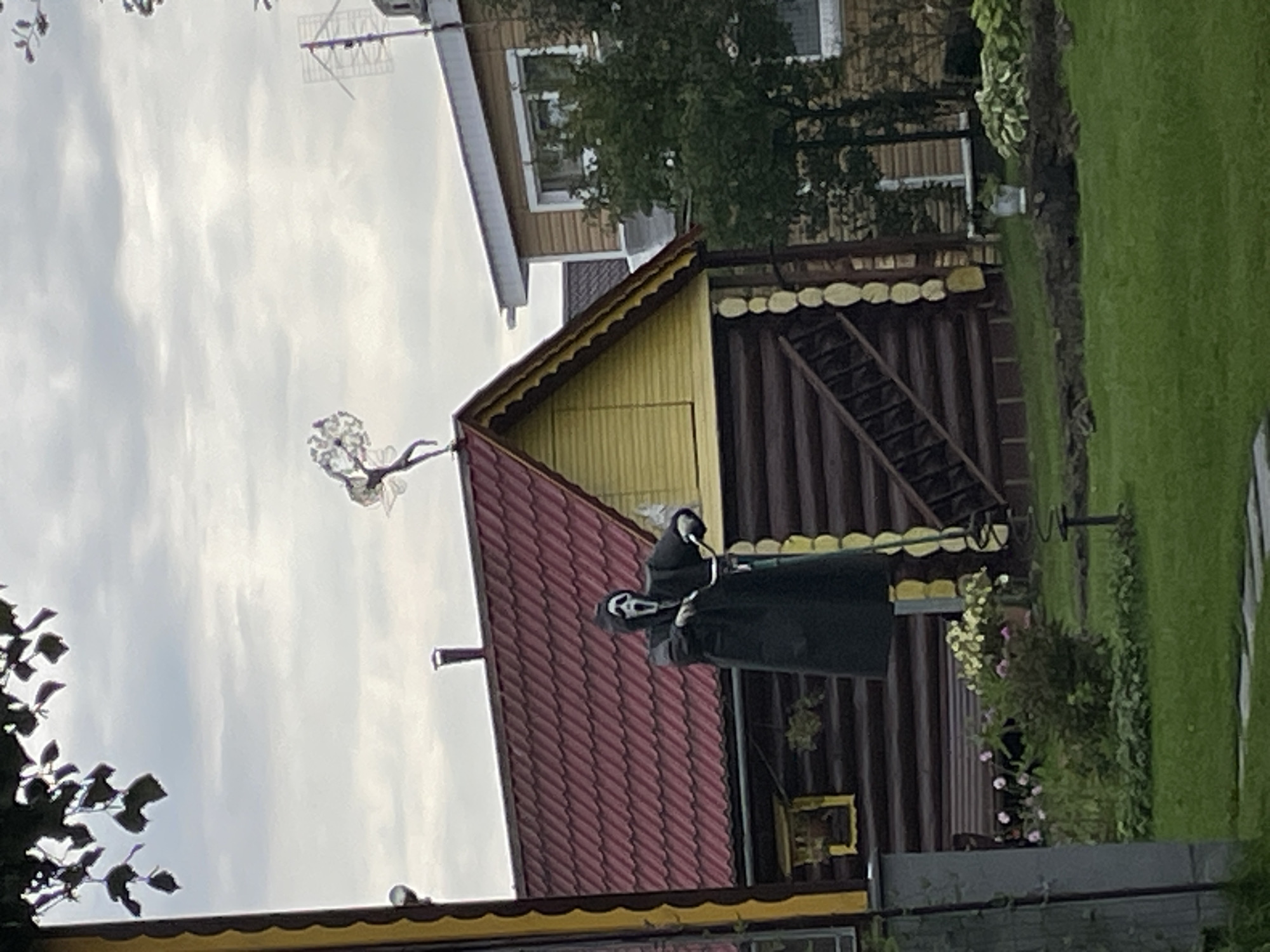 A figure resembling the Ghostface killer from the Scream franchise is positioned outdoors, seemingly riding a bicycle. The figure is dressed in a black robe and mask. Behind the figure, a rustic wooden building with a red roof and yellow trim is visible. Further in the background, another house with light-colored siding and a white roof can be seen, along with mature trees and foliage. The sky is overcast, suggesting a cloudy day. The scene appears to be in a suburban or rural setting, possibly a backyard in Строймаш Снт, Russia.