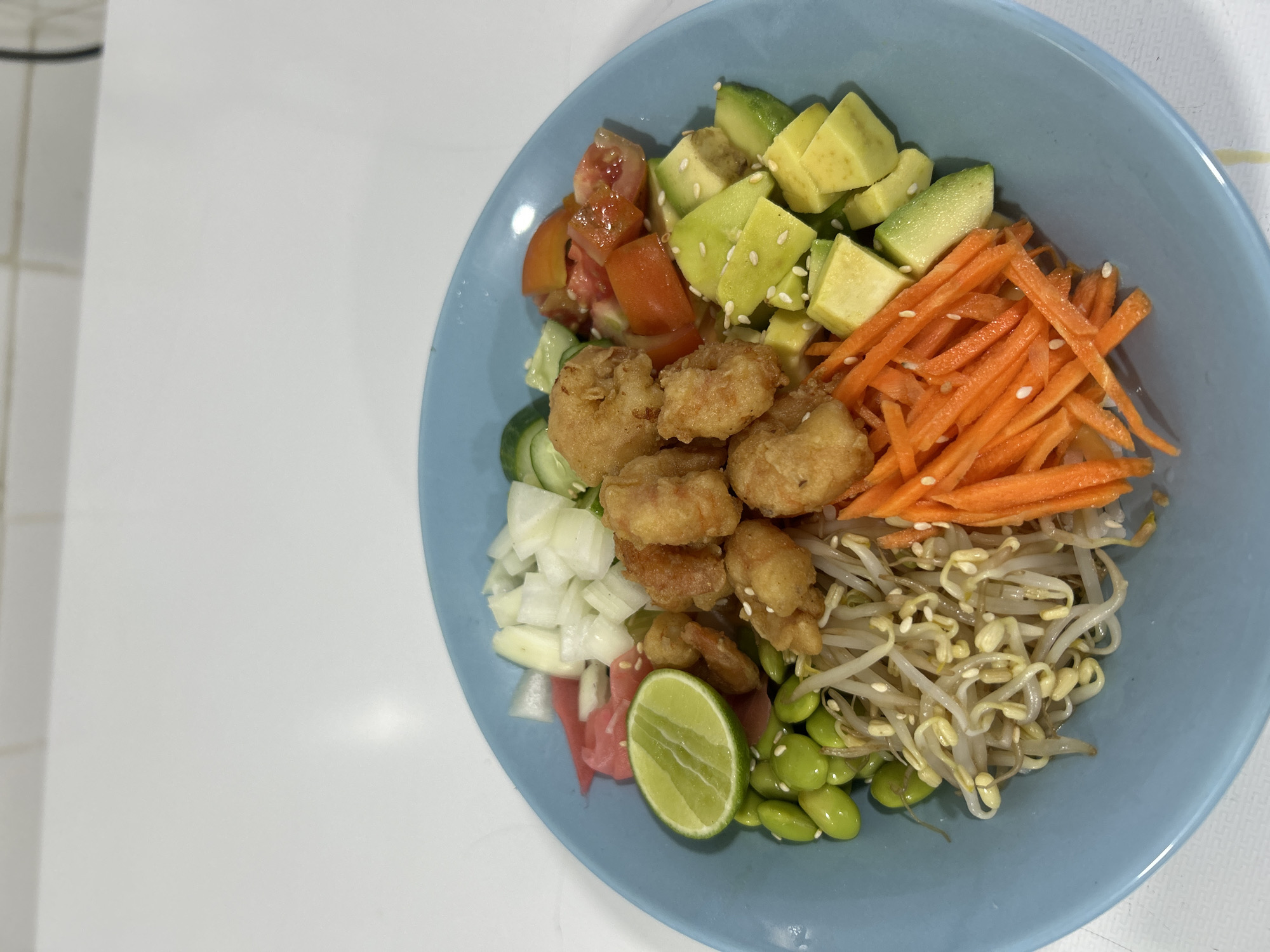 A top-down, eye-level, close-up shot shows a light blue bowl filled with a colorful salad on a white surface. The salad appears to be a composed salad featuring distinct groupings of ingredients. On the upper left are diced tomatoes, followed by cubed avocado sprinkled with sesame seeds. To the right of the avocado are julienned carrots. Continuing clockwise, there are bean sprouts and edamame. Moving left are fried pieces of food, possibly shrimp or chicken, followed by diced white onion, pickled ginger, a lime wedge, and slices of cucumber. The location is unknown and there is no evidence of the city of Senggigi, Indonesia. There are no people or interactions visible. The environment is a clean, flat surface.