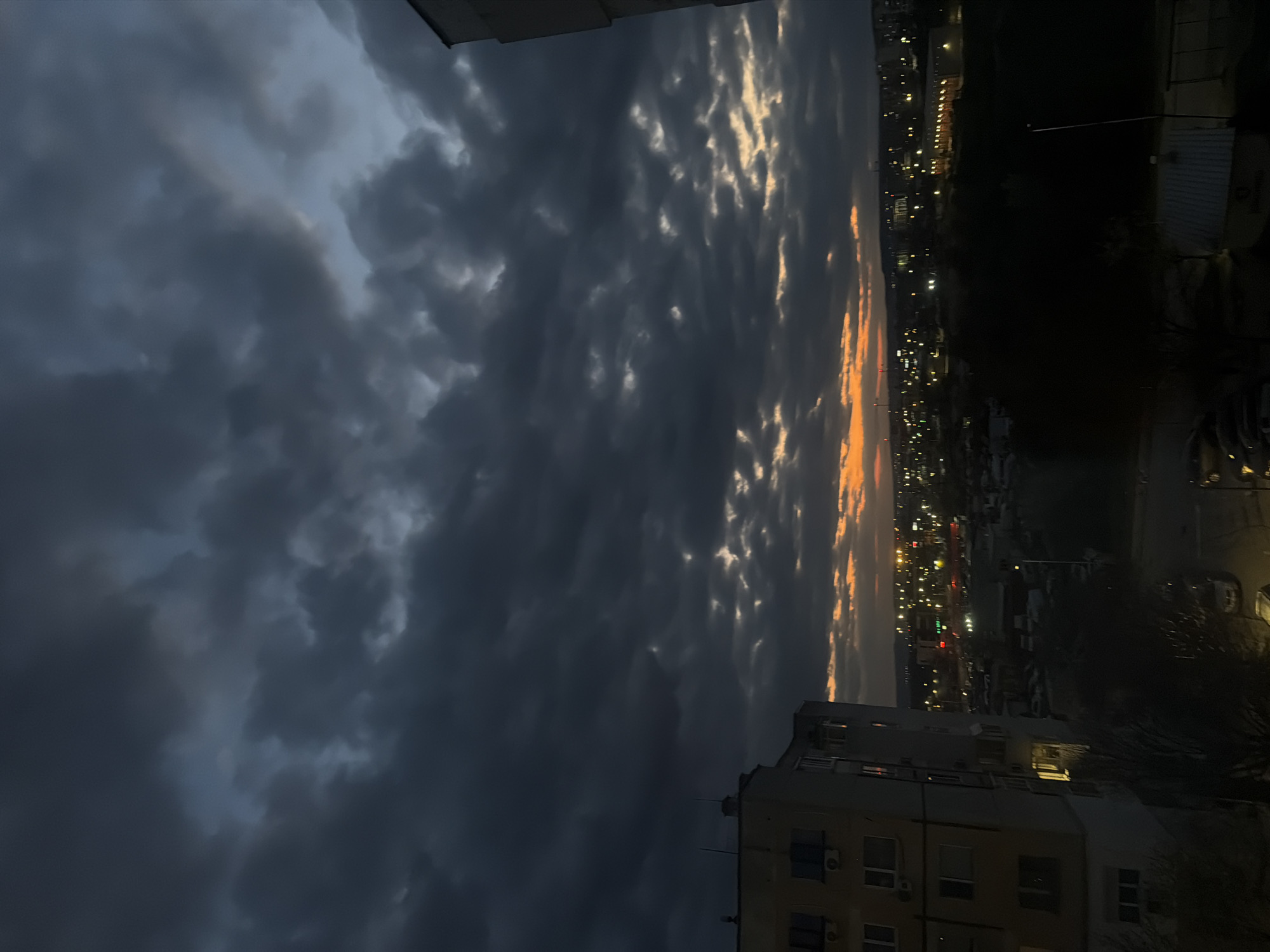 The image, oriented incorrectly as if rotated counter-clockwise, depicts an urban landscape at dusk in Varna, Bulgaria. The dominant feature is the sky, filled with heavy, dark, and turbulent cloud formations, suggesting an impending storm or a recent weather event. Patches of these clouds are illuminated by the setting sun, revealing intricate, wavelike textures, possibly indicative of undulatus asperatus clouds, with hues of golden yellow and orange contrasting sharply against the dark grays and blues of the other clouds.
On the horizon, a distant city line stretches across the mid-ground, marked by numerous twinkling city lights that glow warmly against the dimming sky. A prominent multi-story building, likely an apartment block with a typical Eastern European architectural style, occupies the lower right corner of the frame. Its windows show interior lights, indicating habitation.
In the darker areas of the mid-ground, outlines of trees or vegetation are faintly visible, and what appear to be parked cars can be discerned. No specific activity or event is taking place; the image captures a static, dramatic moment at twilight. There is no visible text. The overall mood is one of an urban evening under a powerful and visually striking sky.